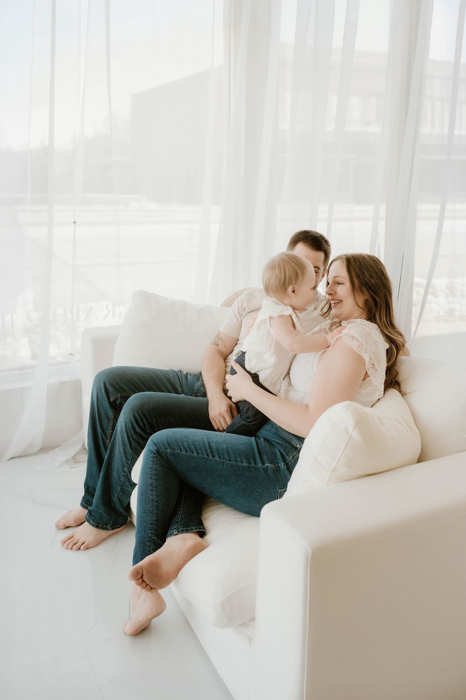 Happy young family with baby sitting on white sofa near bright windows in a cozy living room.