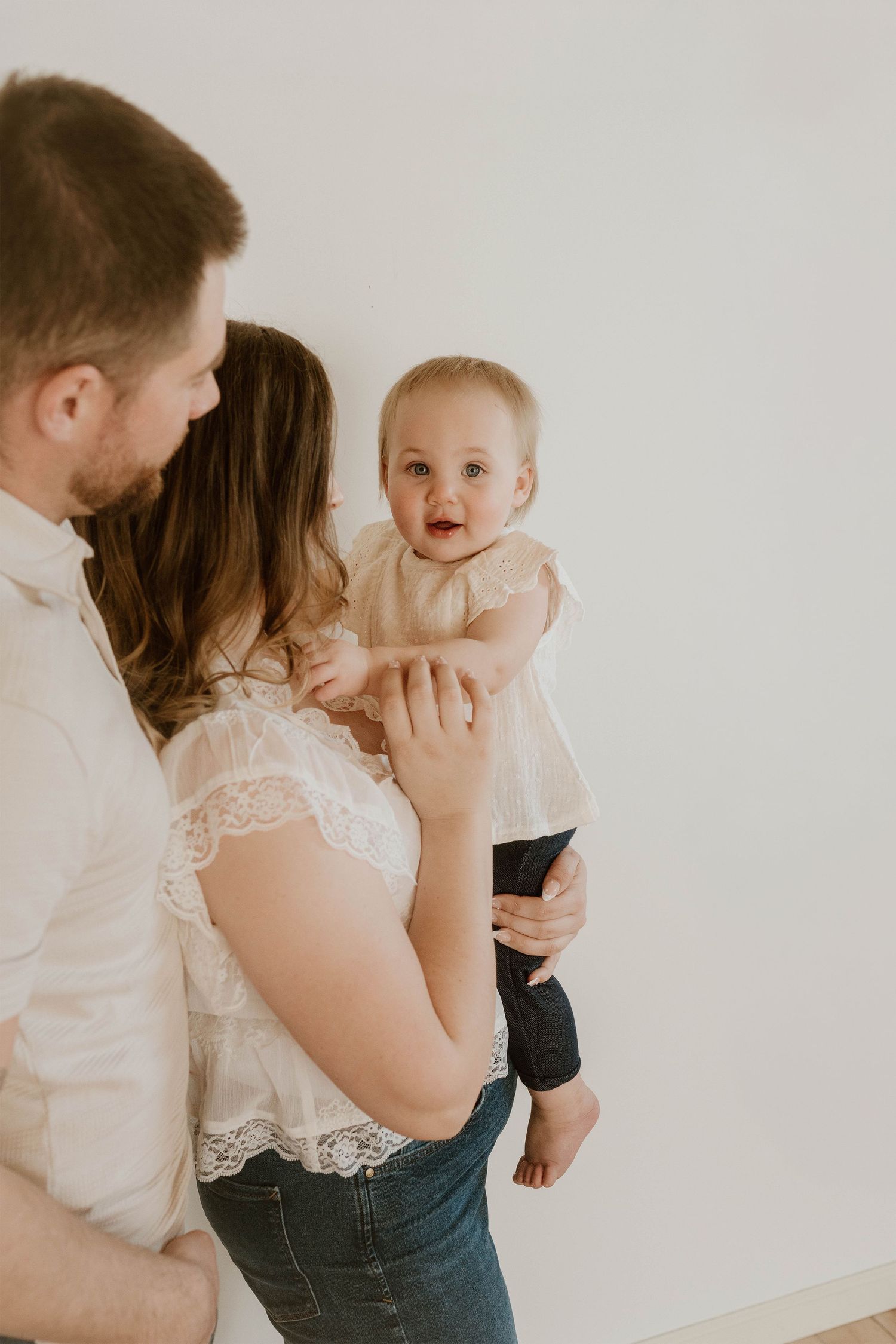 A couple holds their smiling baby girl in a bright, minimalist studio during a family portrait session.
