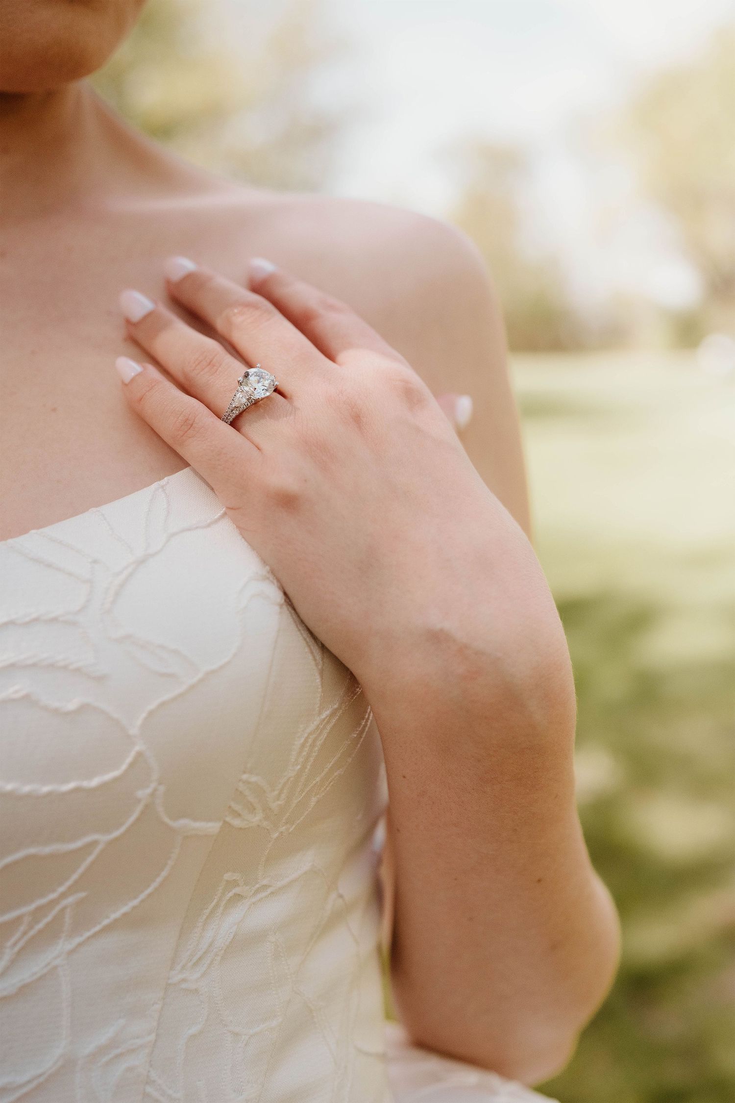 Bride in strapless white gown with hand on chest showcasing a sparkling diamond engagement ring outdoors.