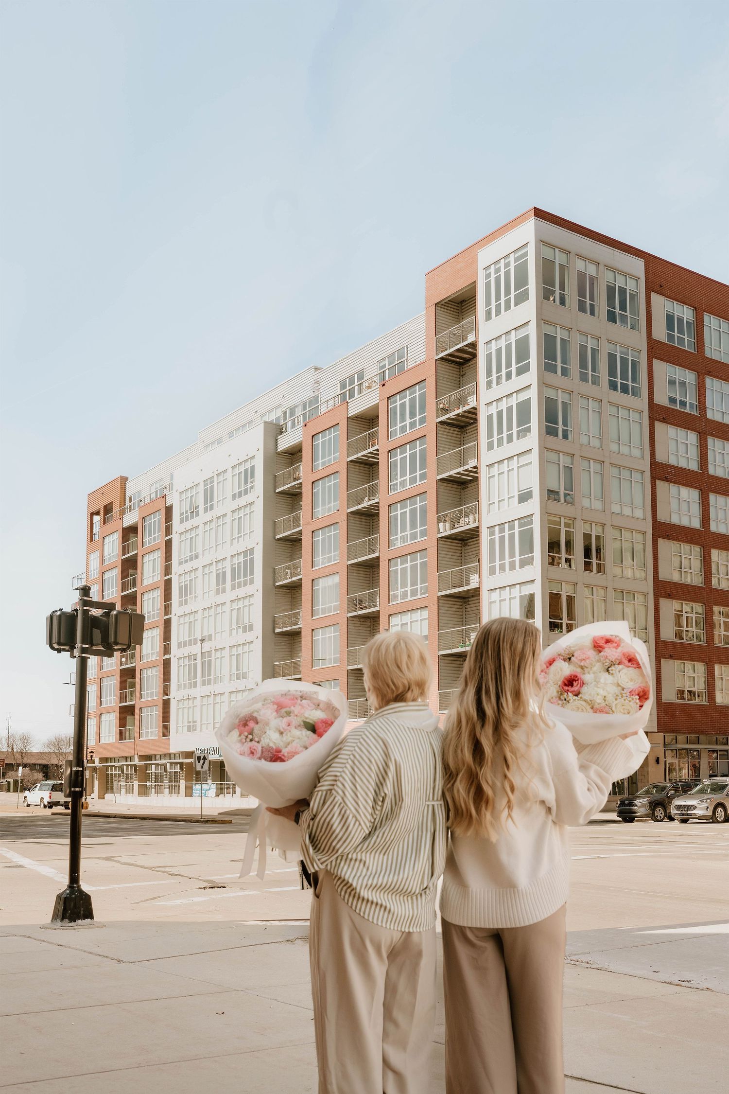 Two women holding large pink and white flower bouquets stand on a city street corner with a modern brick building behind them.