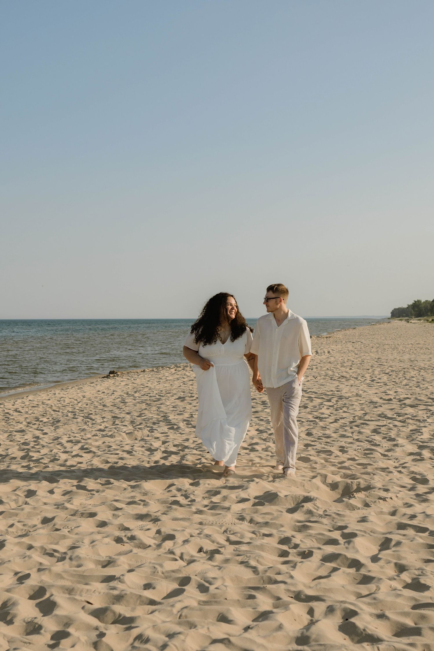 Couple in white outfits walking hand in hand on a sandy beach with calm ocean waters in the background.
