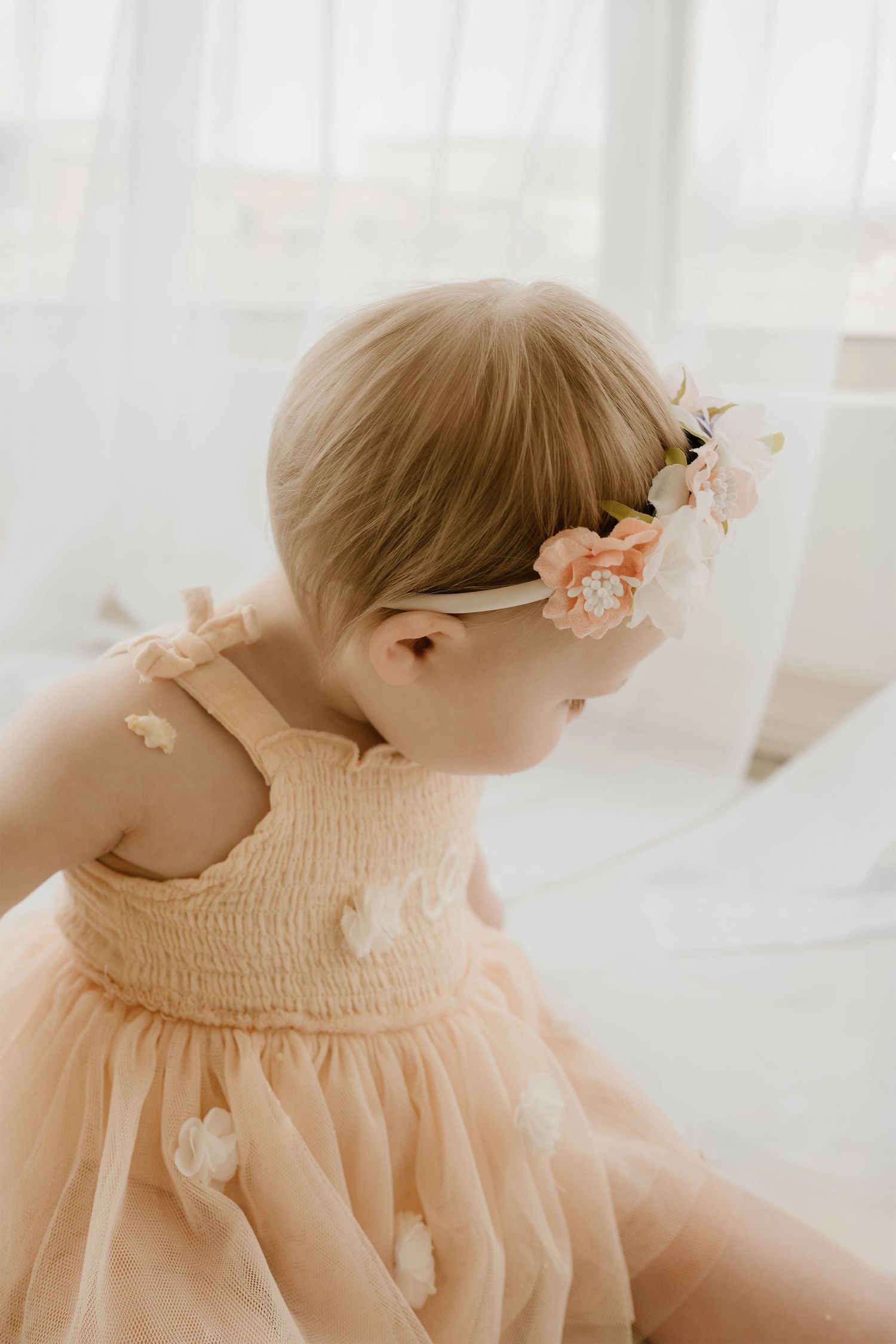 Baby girl in peach smocked tulle dress with floral headband, soft natural light portrait photography.