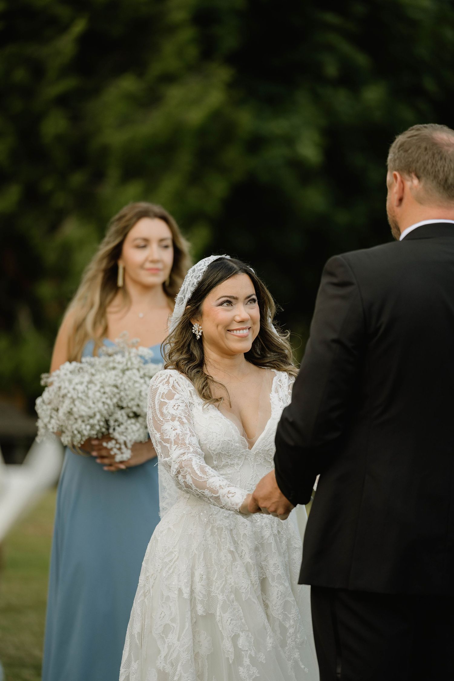 Bride in lace gown smiles at groom during outdoor wedding ceremony, bridesmaid in blue holds white bouquet behind her.