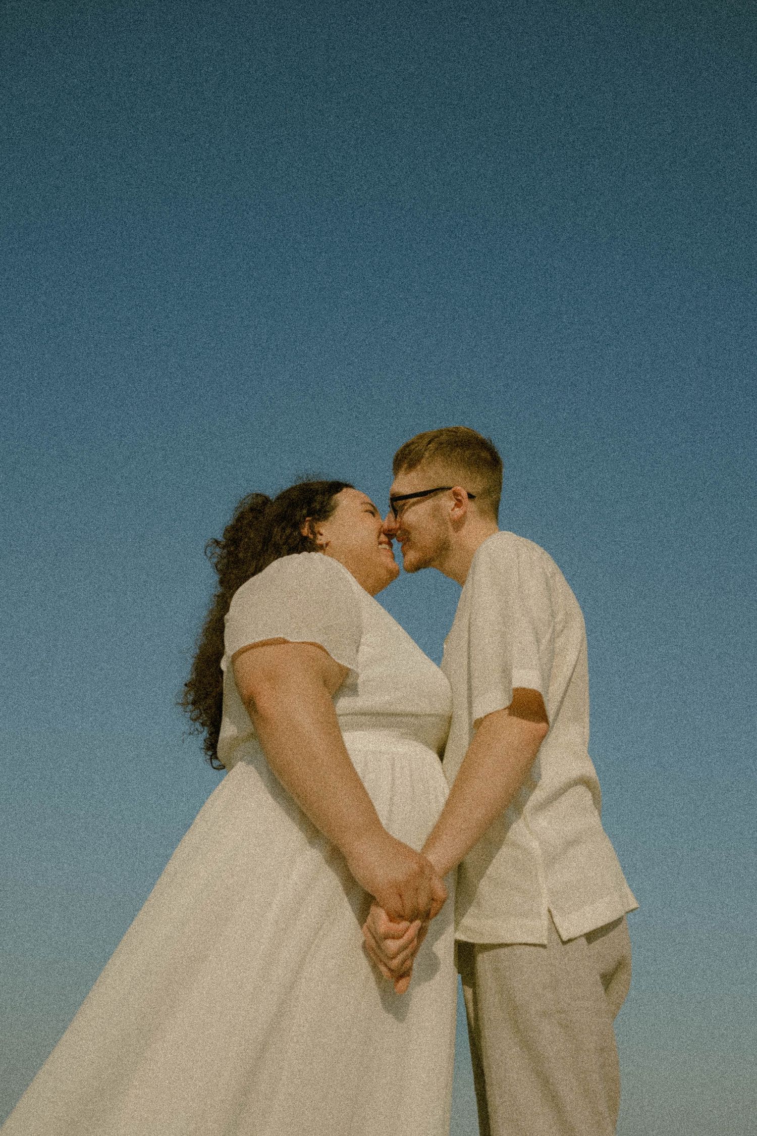 A couple in white outfits holds hands and leans in for a kiss against a clear blue sky.
