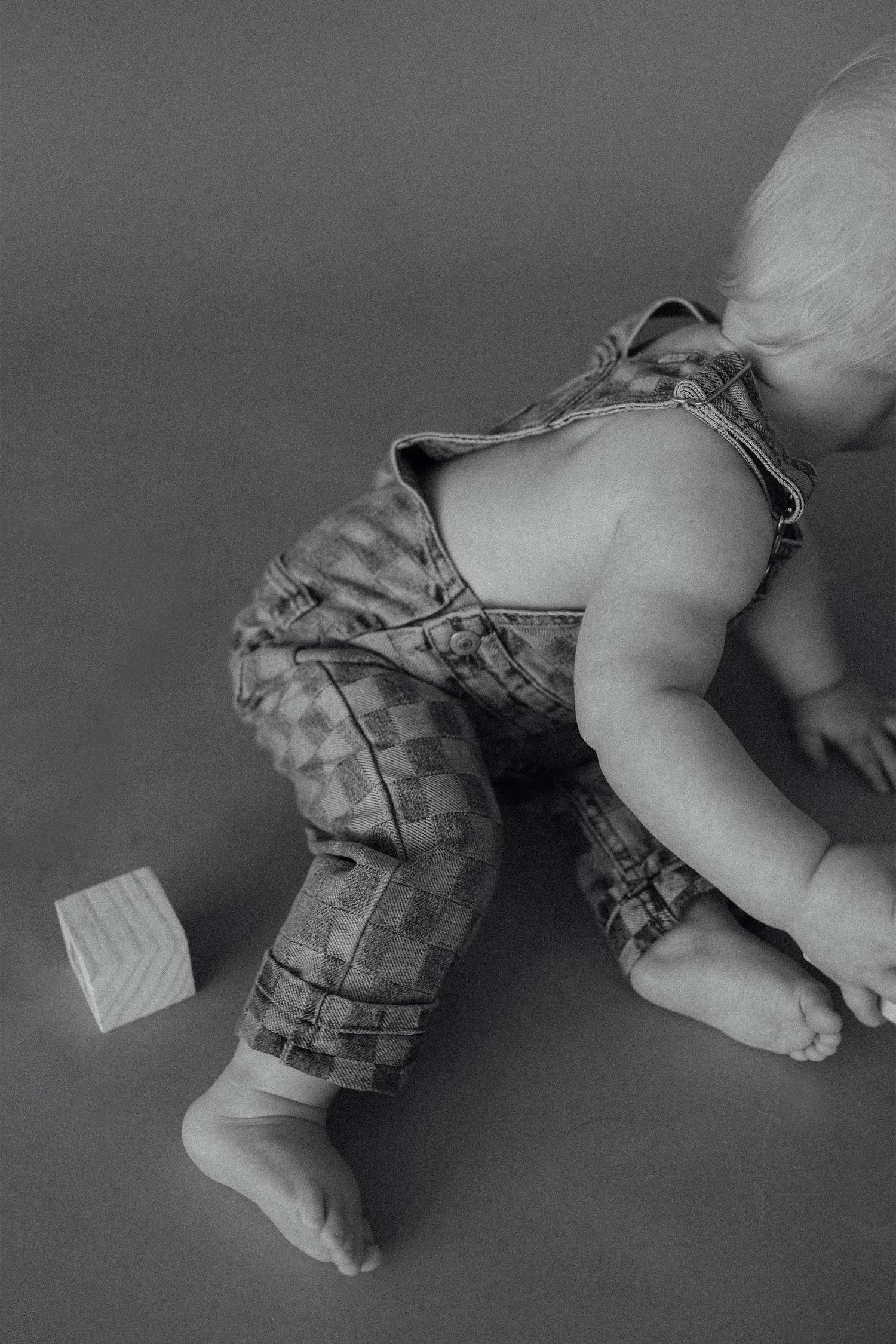 Black and white photo of a baby in checkered overalls playing on the floor next to a wooden block toy.