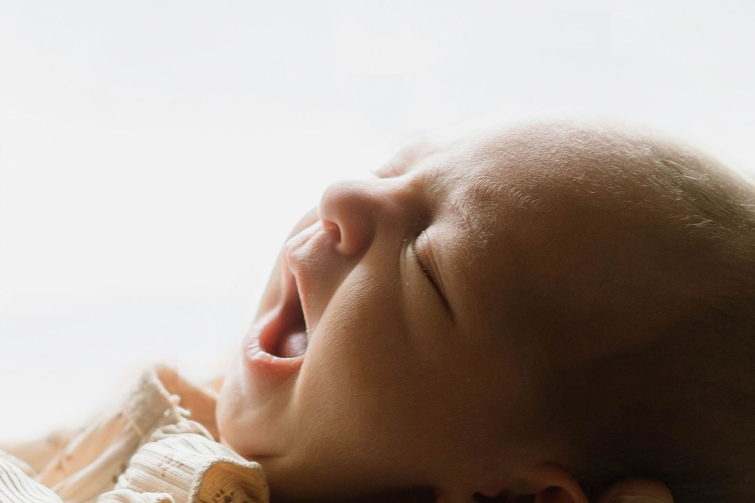 Close-up of a newborn baby crying with eyes closed, lying on a soft blanket against a white background.