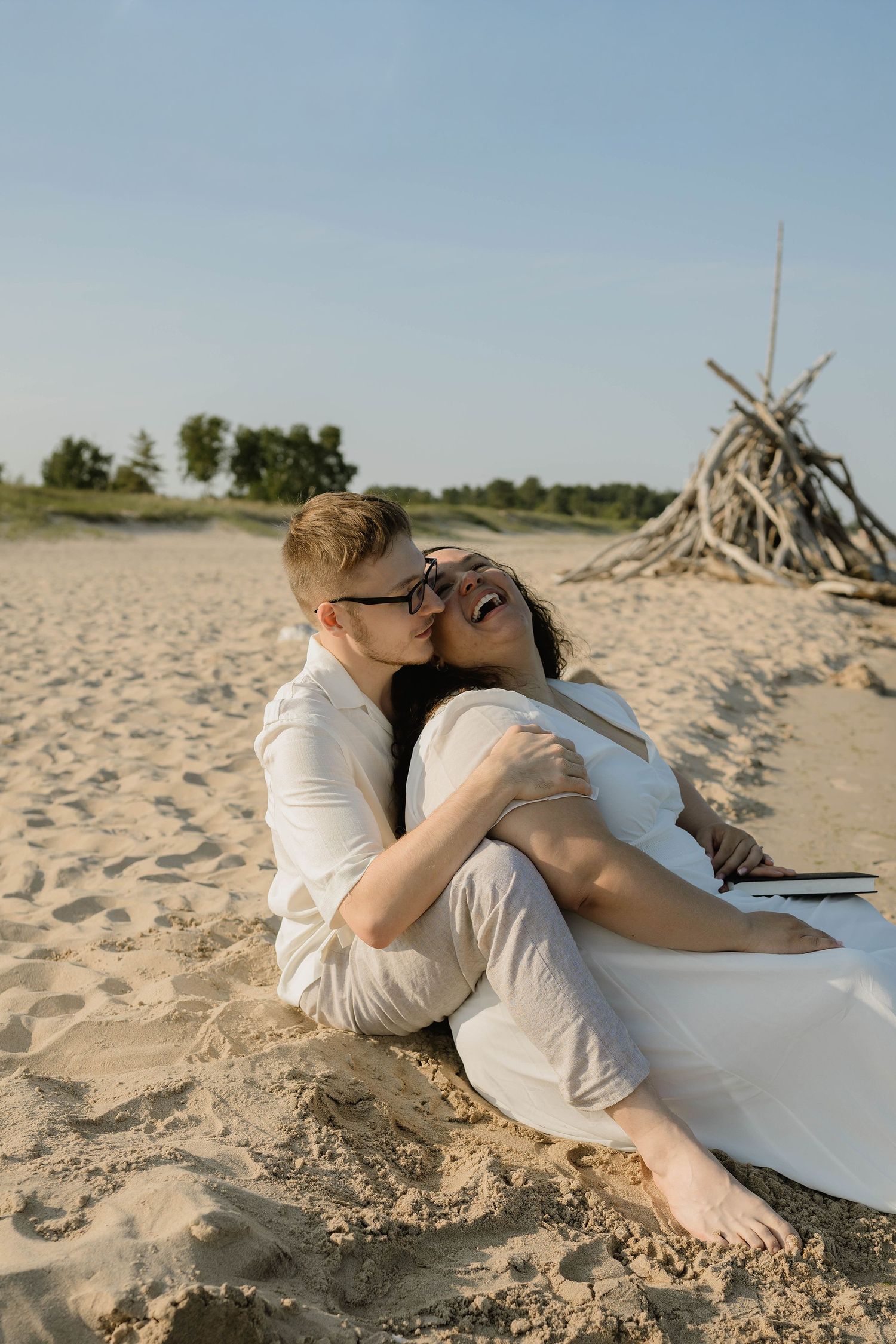 A couple in white sits on sandy beach, laughing and embracing near a driftwood structure.