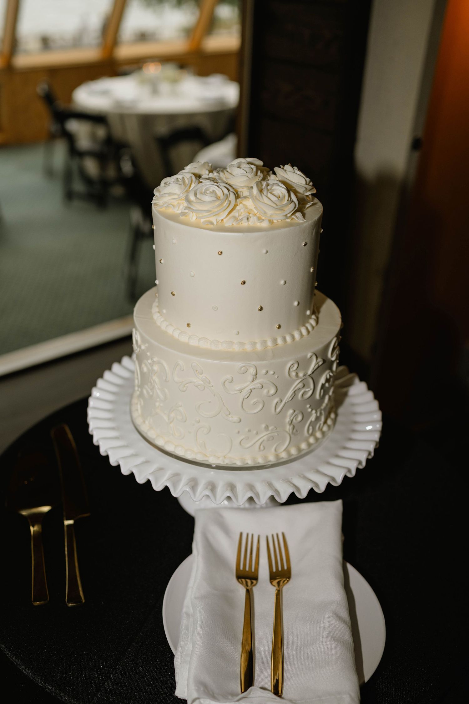 Elegant two-tier white wedding cake with floral topper, scroll details, and gold dot accents displayed on a ruffled stand.