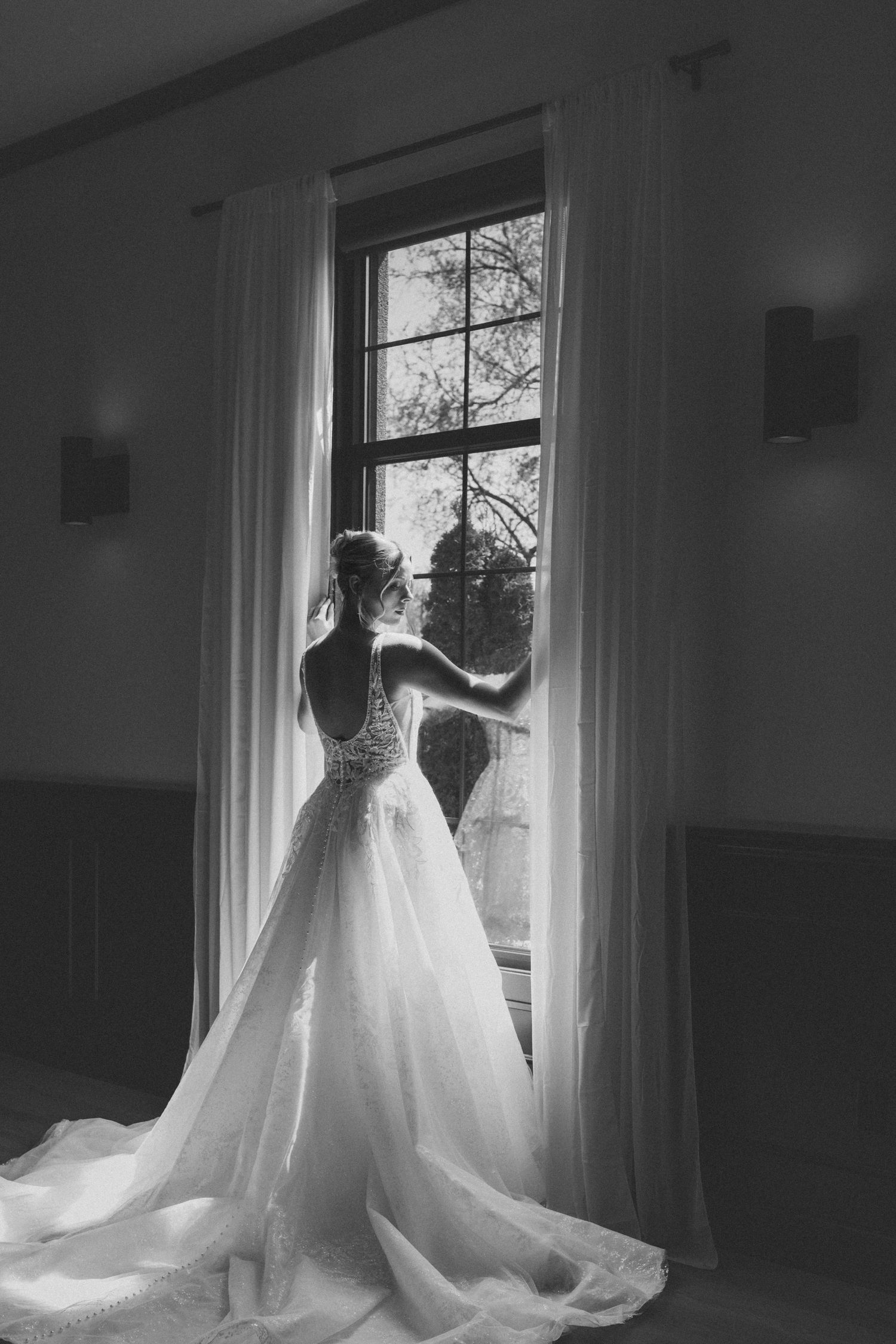 Black and white photo of a bride in a flowing wedding gown standing before a tall window with curtains.