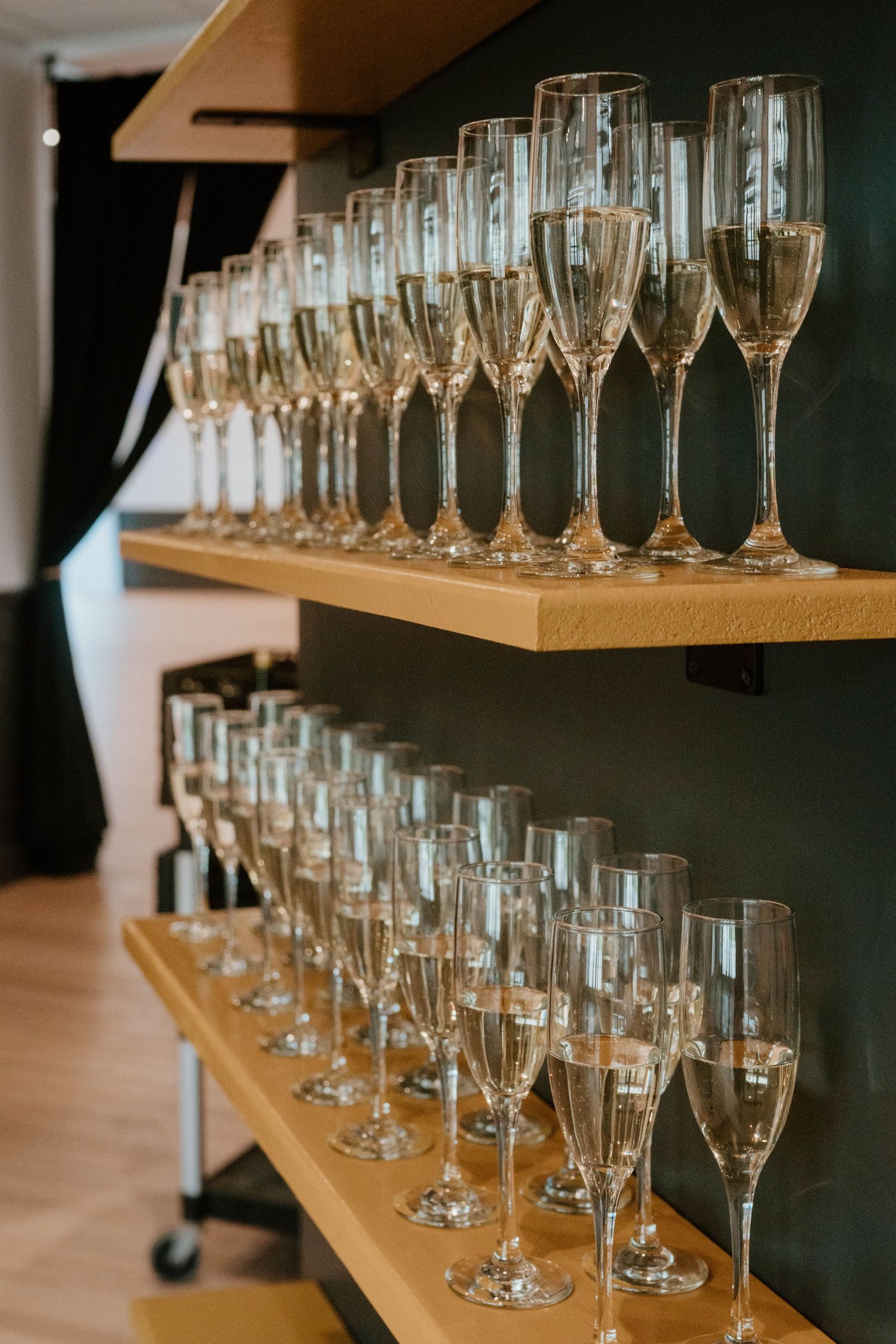 Rows of elegant champagne flute glasses neatly arranged on wooden shelves against a dark green wall.