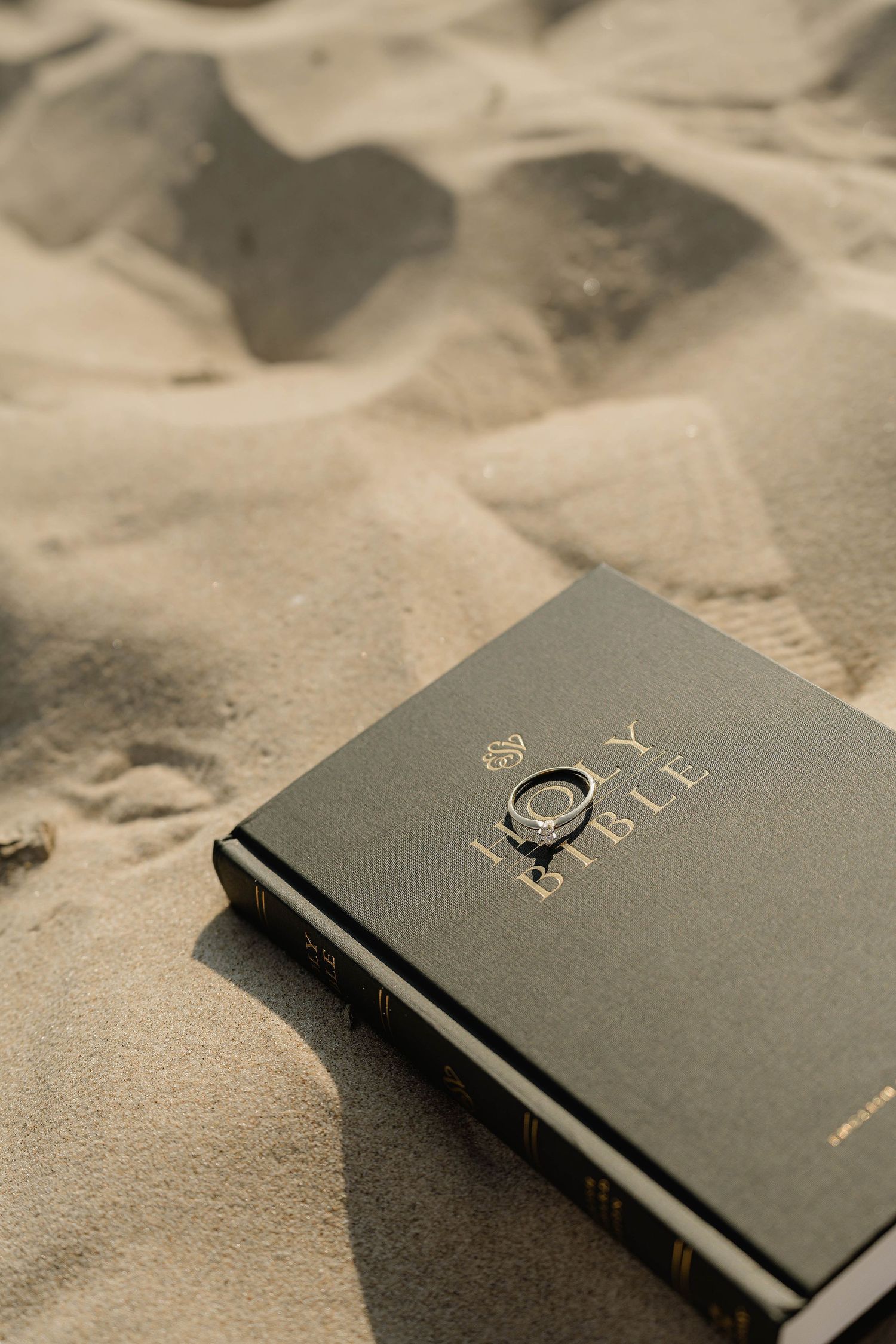 A Holy Bible with a ring resting on top, placed on sandy beach in warm sunlight.