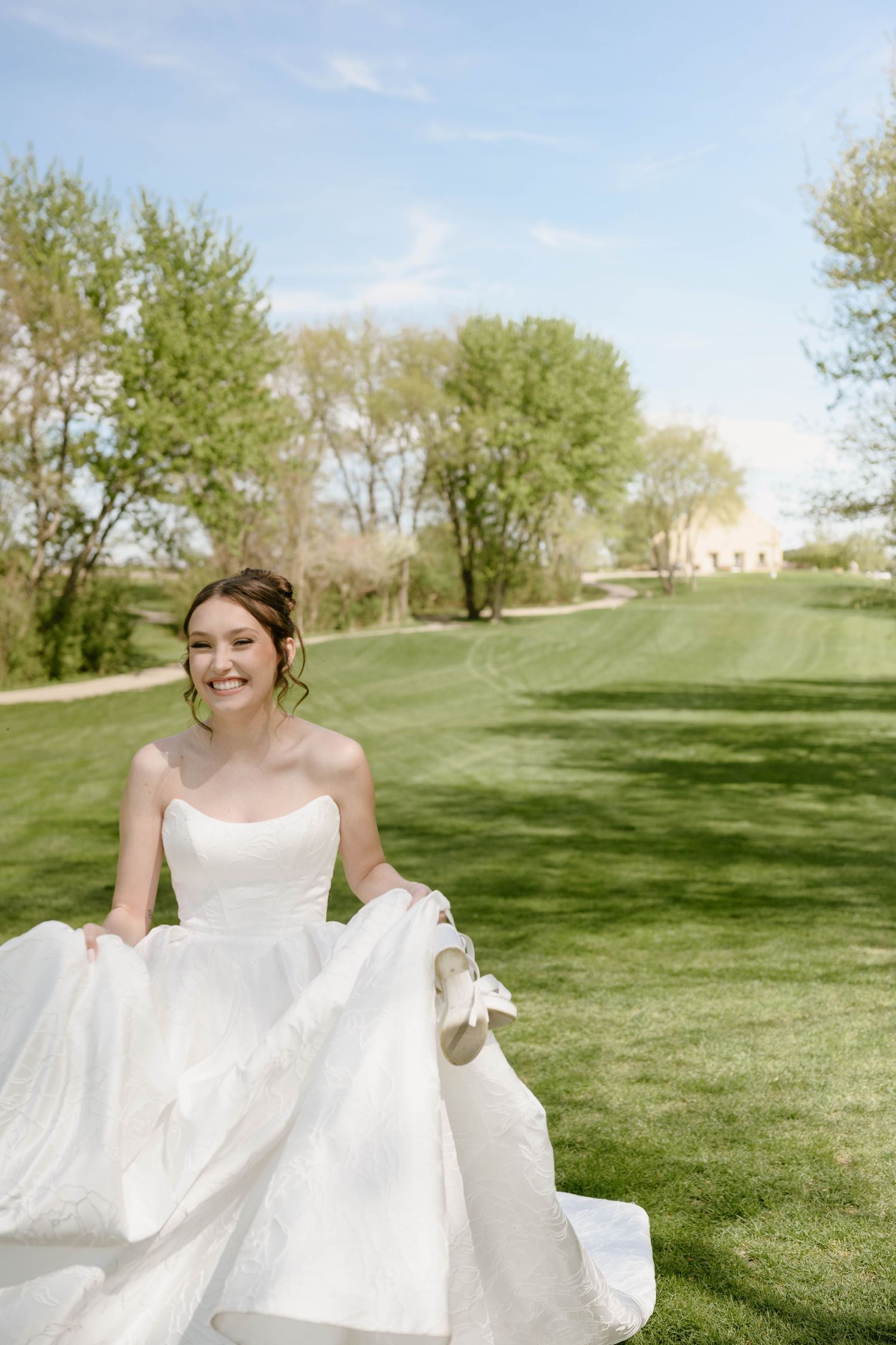 Smiling bride in a strapless white ball gown holding her skirt on a lush green golf course on a sunny day.