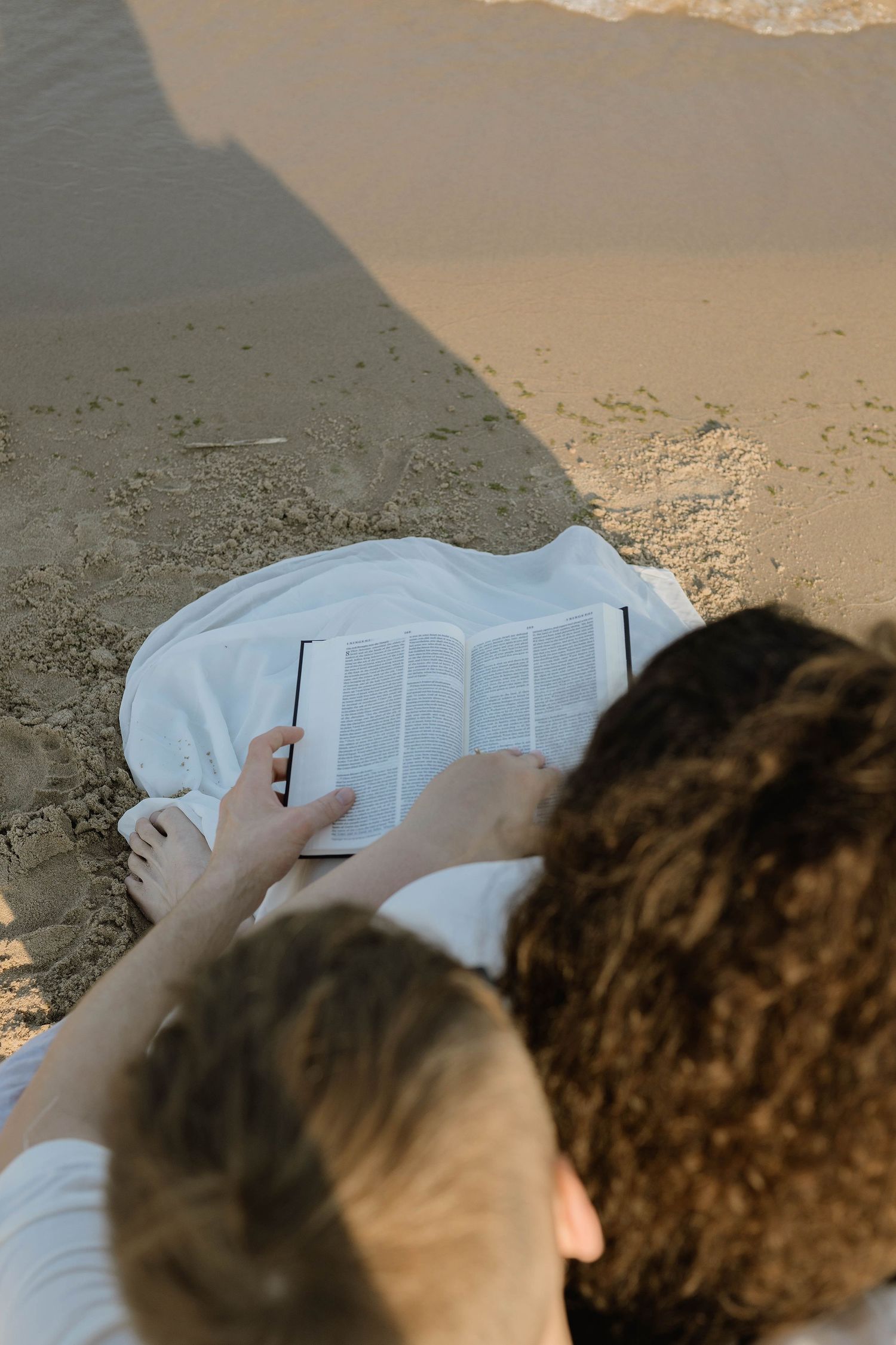 Two people read an open book together while sitting on a white blanket at the beach shoreline.