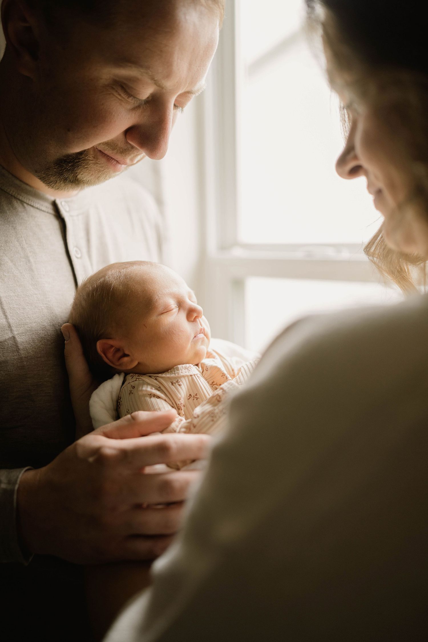 Smiling parents tenderly holding their sleeping newborn baby near a bright window.