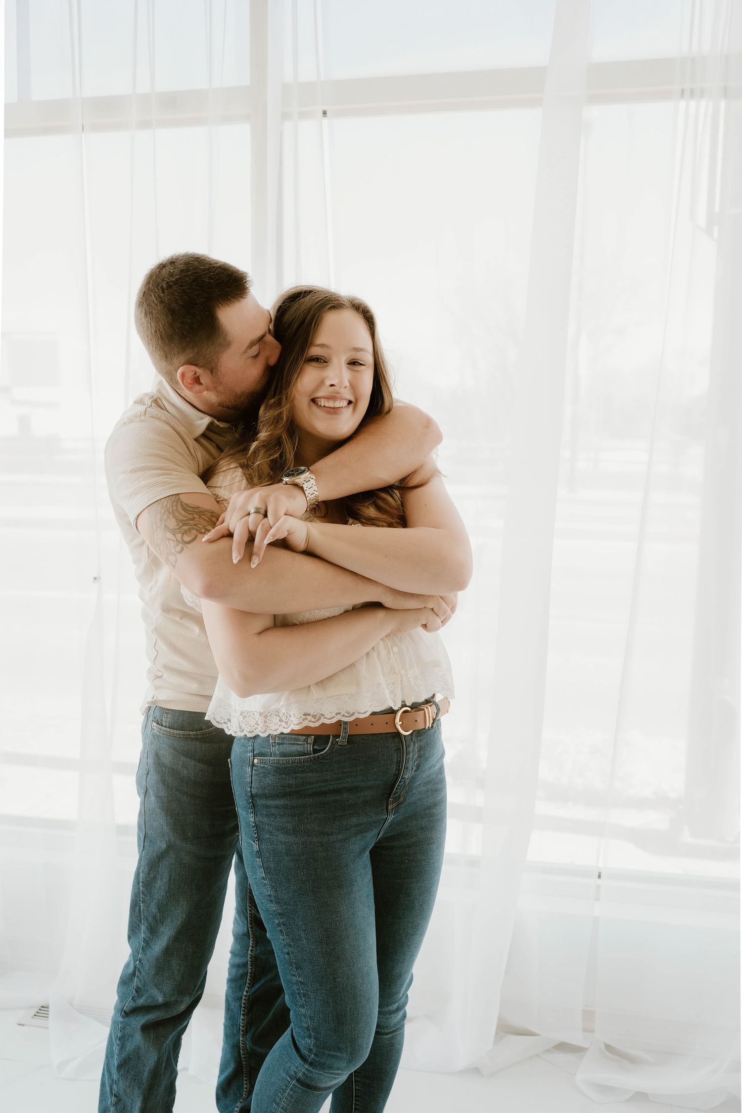 A happy couple embraces and shares a tender kiss in a bright, light-filled white studio setting.