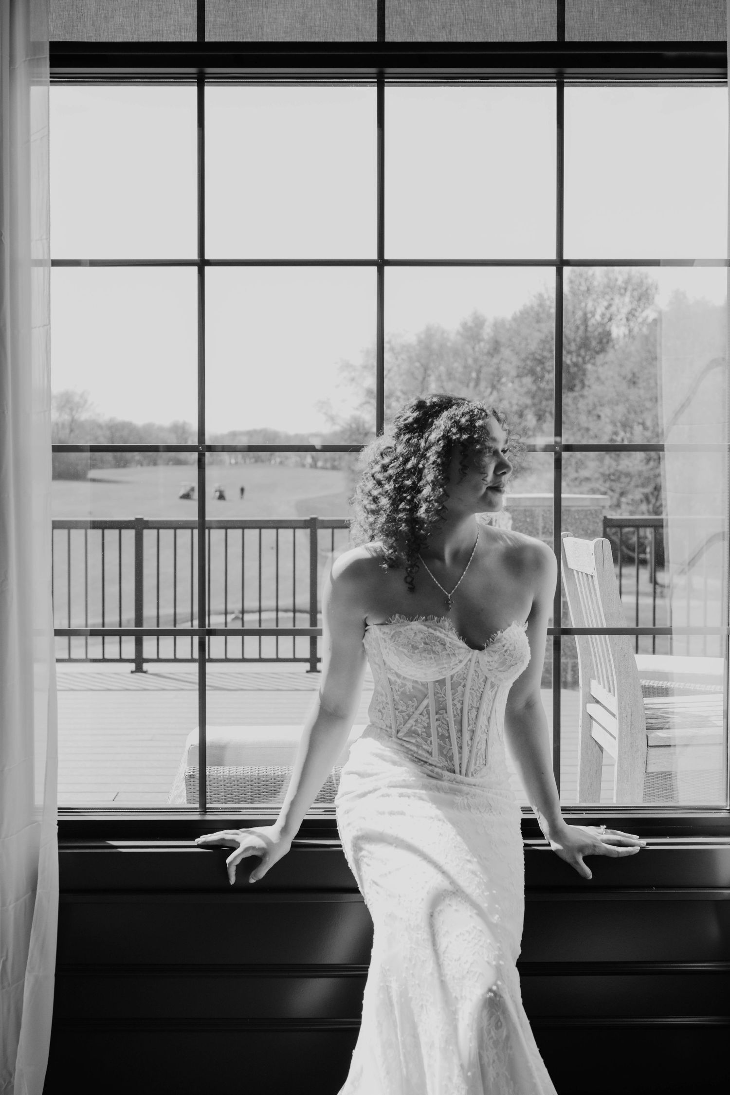 Black and white photo of a bride in a lace wedding gown gazing out a large window on her wedding day.