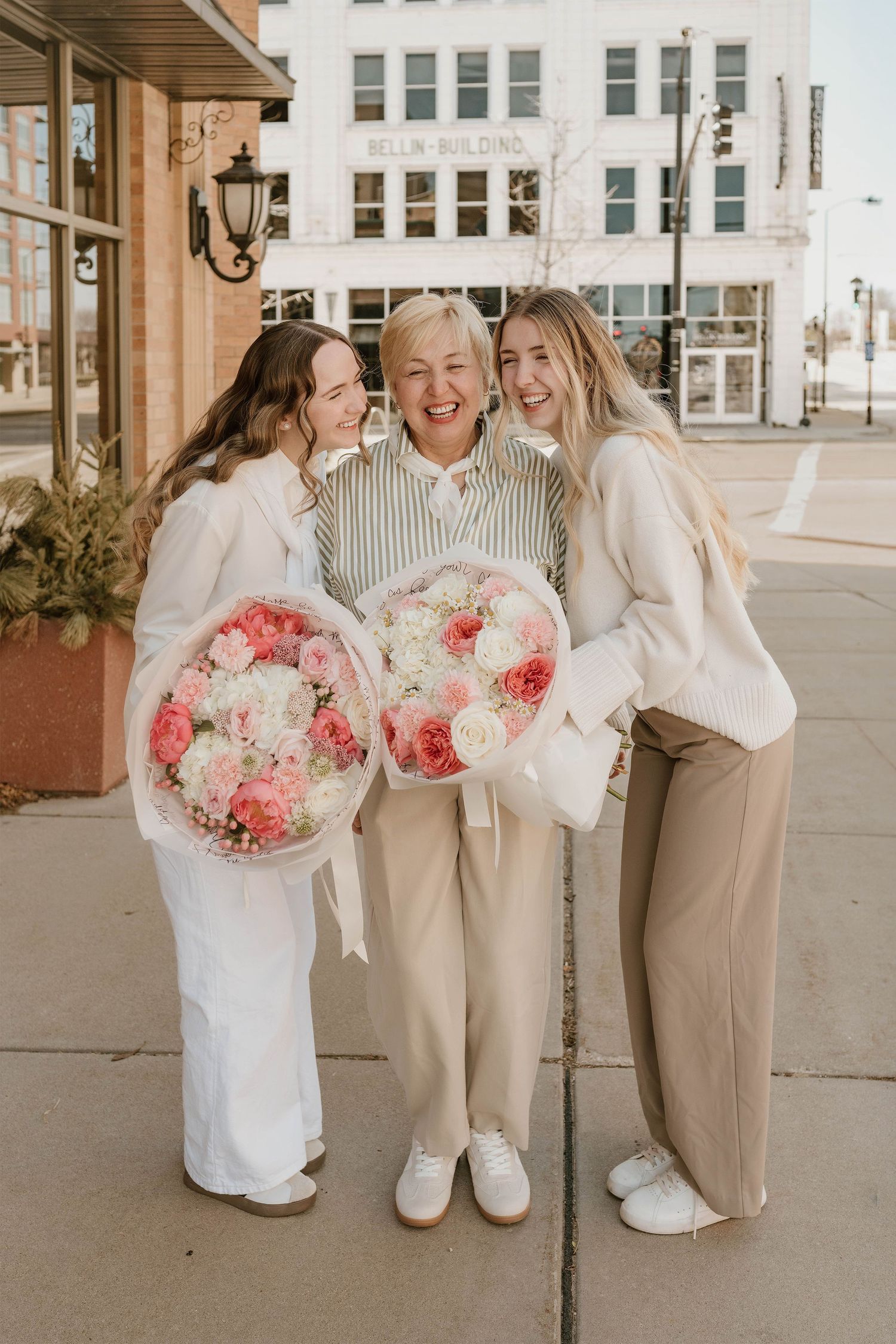 Three women smile outdoors holding large pink and white floral bouquets near a brick building.