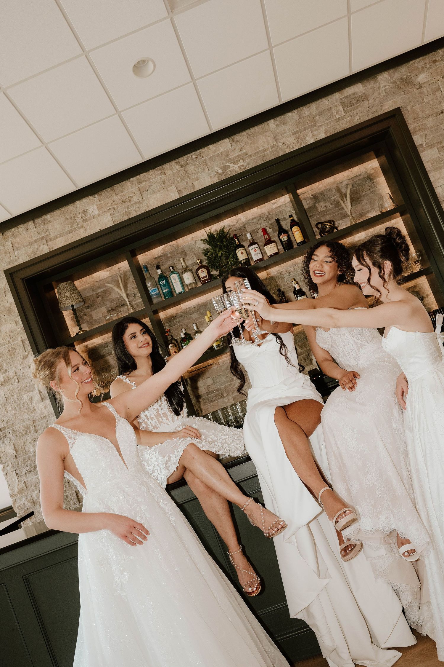 Four bridesmaids in white gowns toasting champagne glasses at a bar with a stone wall backdrop.