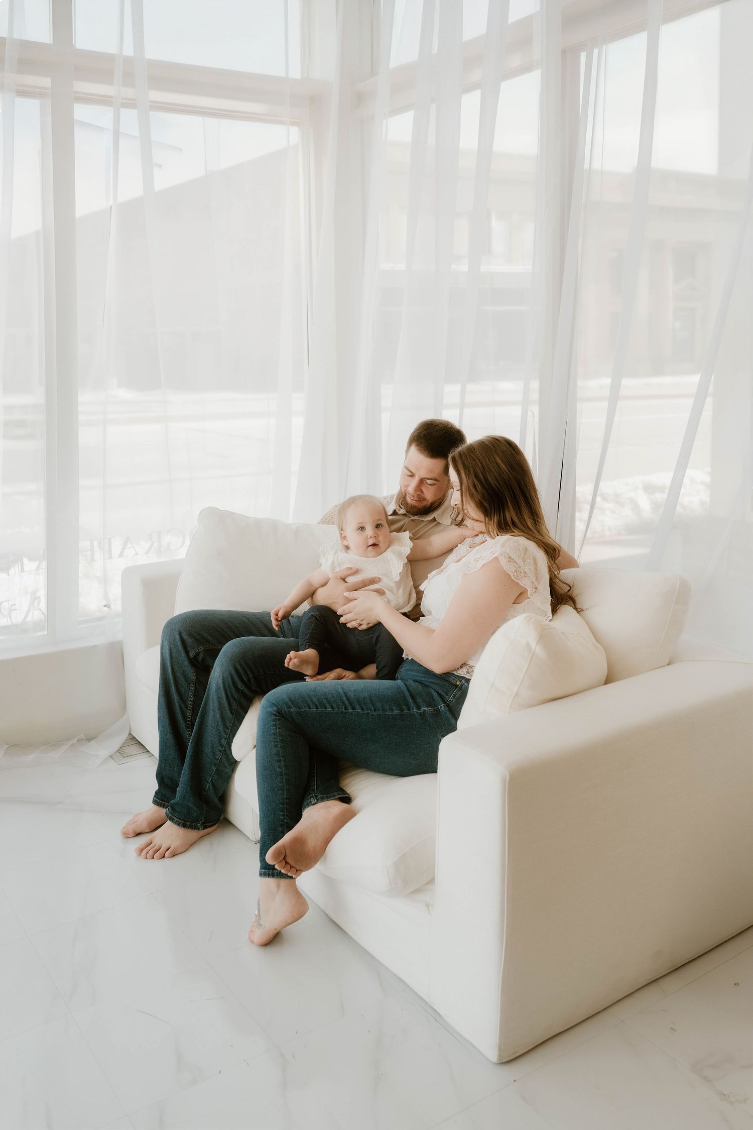 A young family with a baby sits together on a white sofa in a bright, airy living room with large windows.