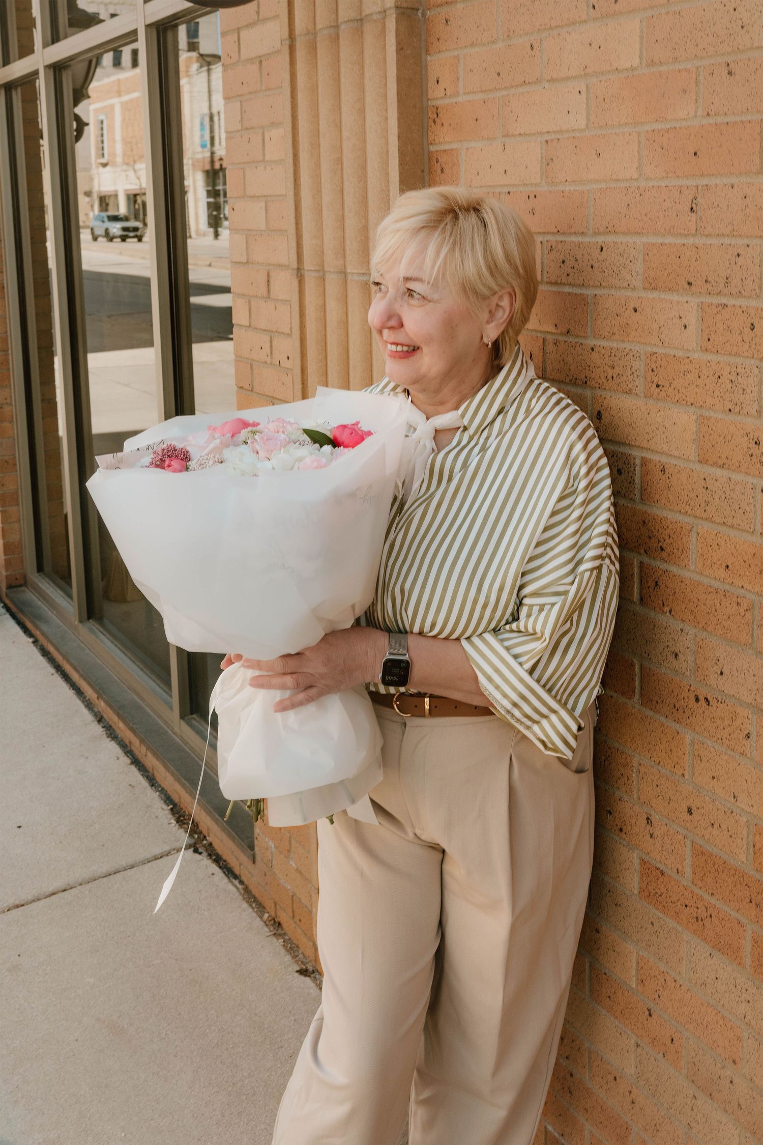 Smiling blonde woman in striped shirt holding large pink floral bouquet wrapped in white paper leaning against brick wall.