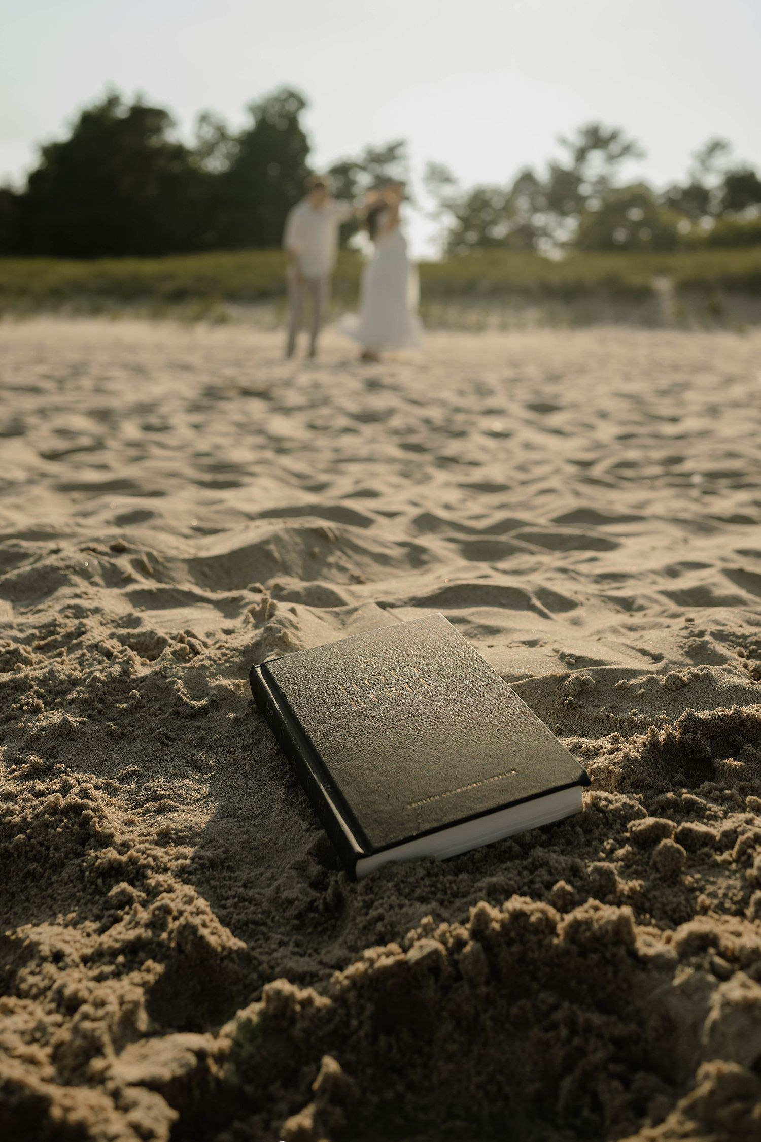 A Holy Bible rests in the sand on a beach with a couple embracing in soft focus in the background.