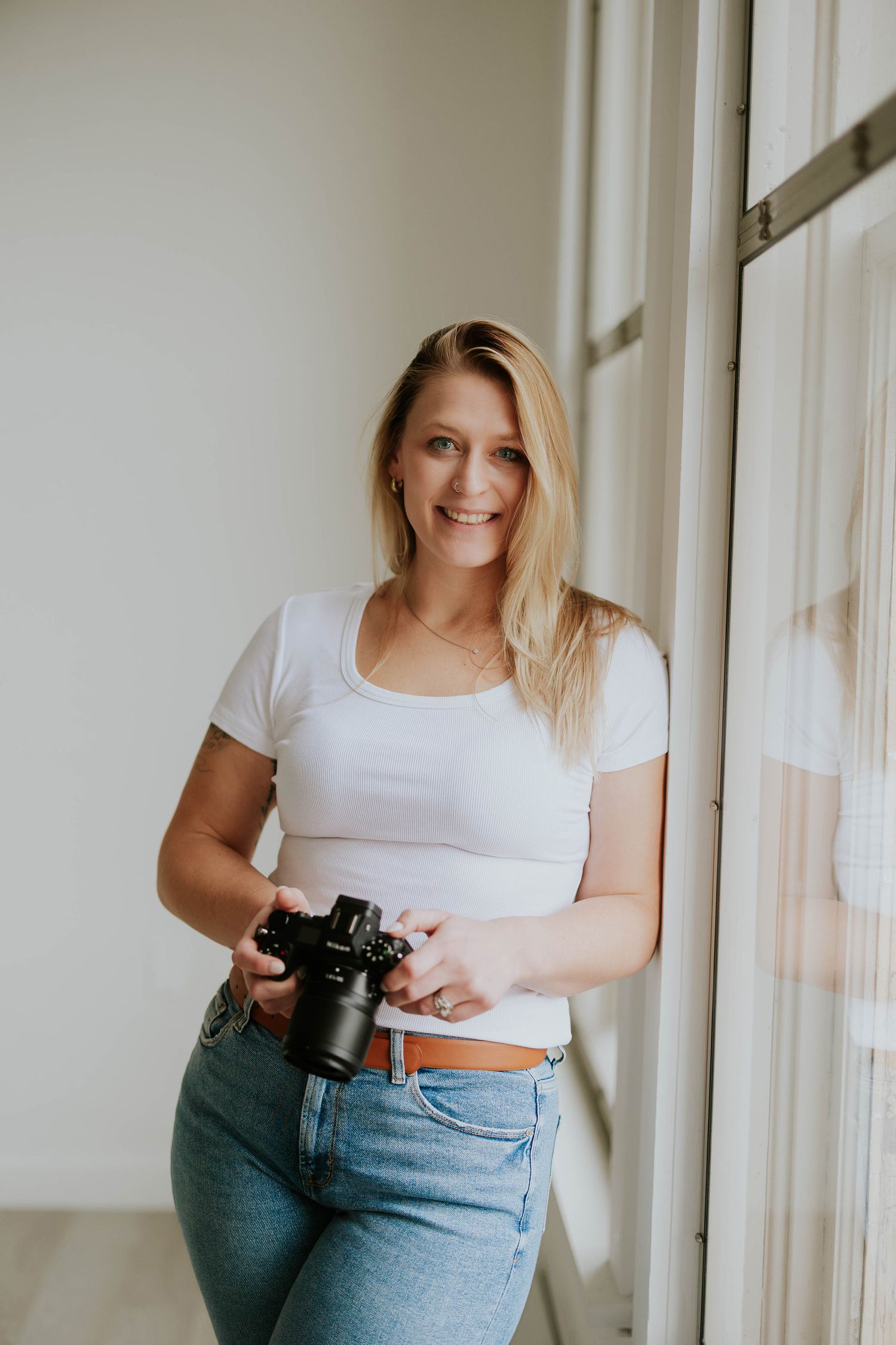 A person in a white t-shirt and jeans holds a camera while standing near bright windows in a minimalist setting.