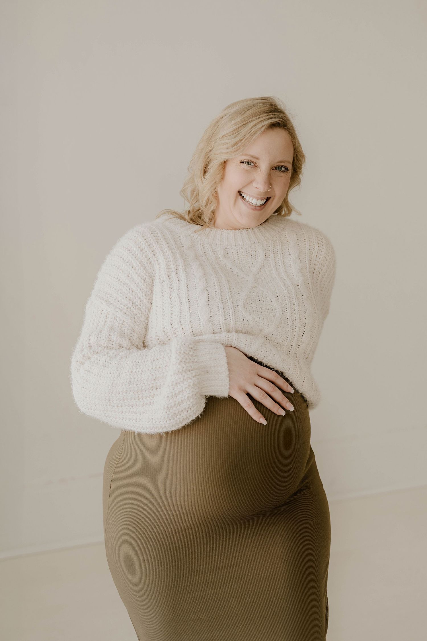 Maternity photo series showing person in cream sweater and olive skirt against neutral backdrop in artistic poses.