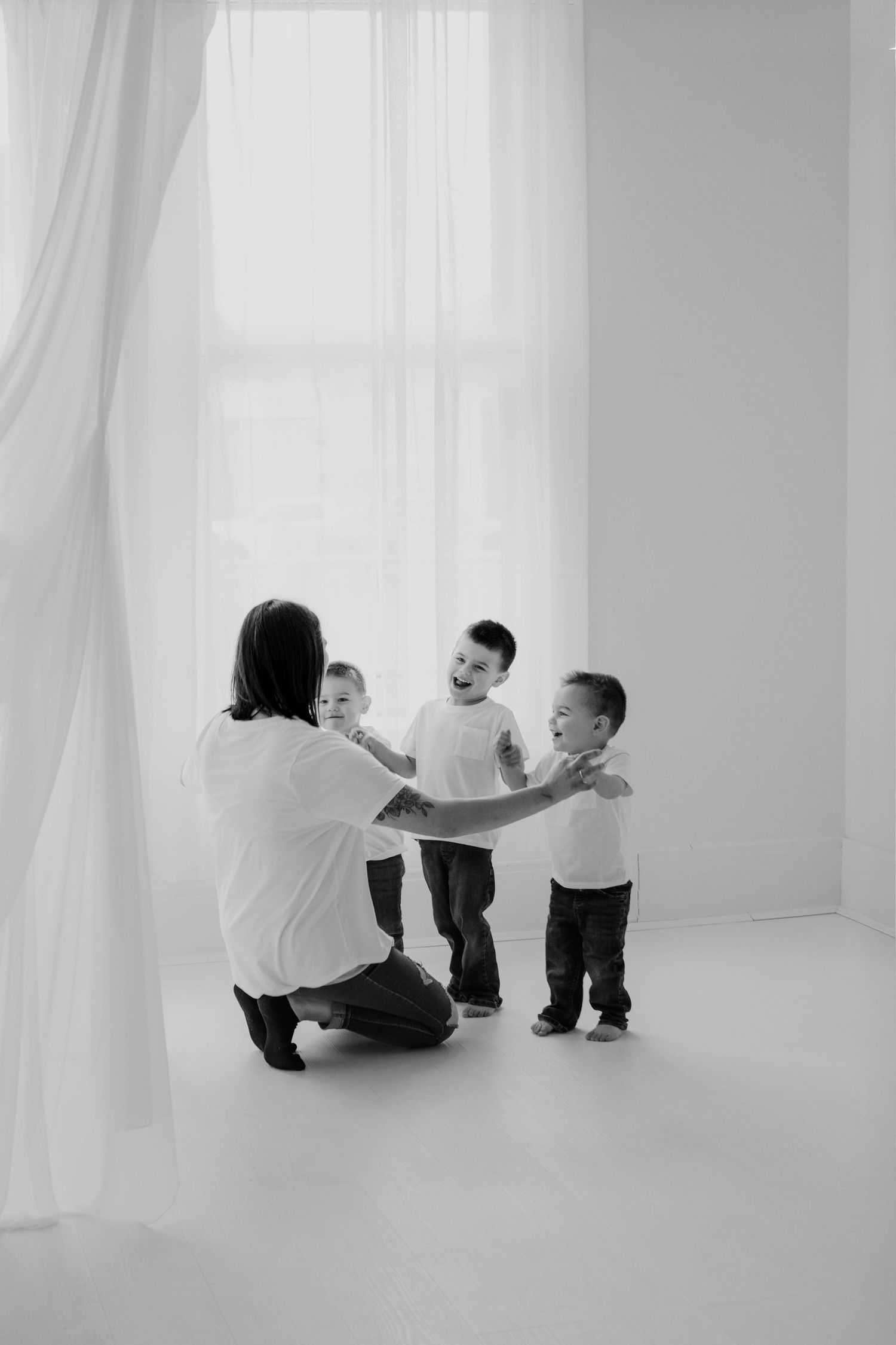 A black and white photo series showing playful interaction between adults and toddlers in a bright white room with curtains.