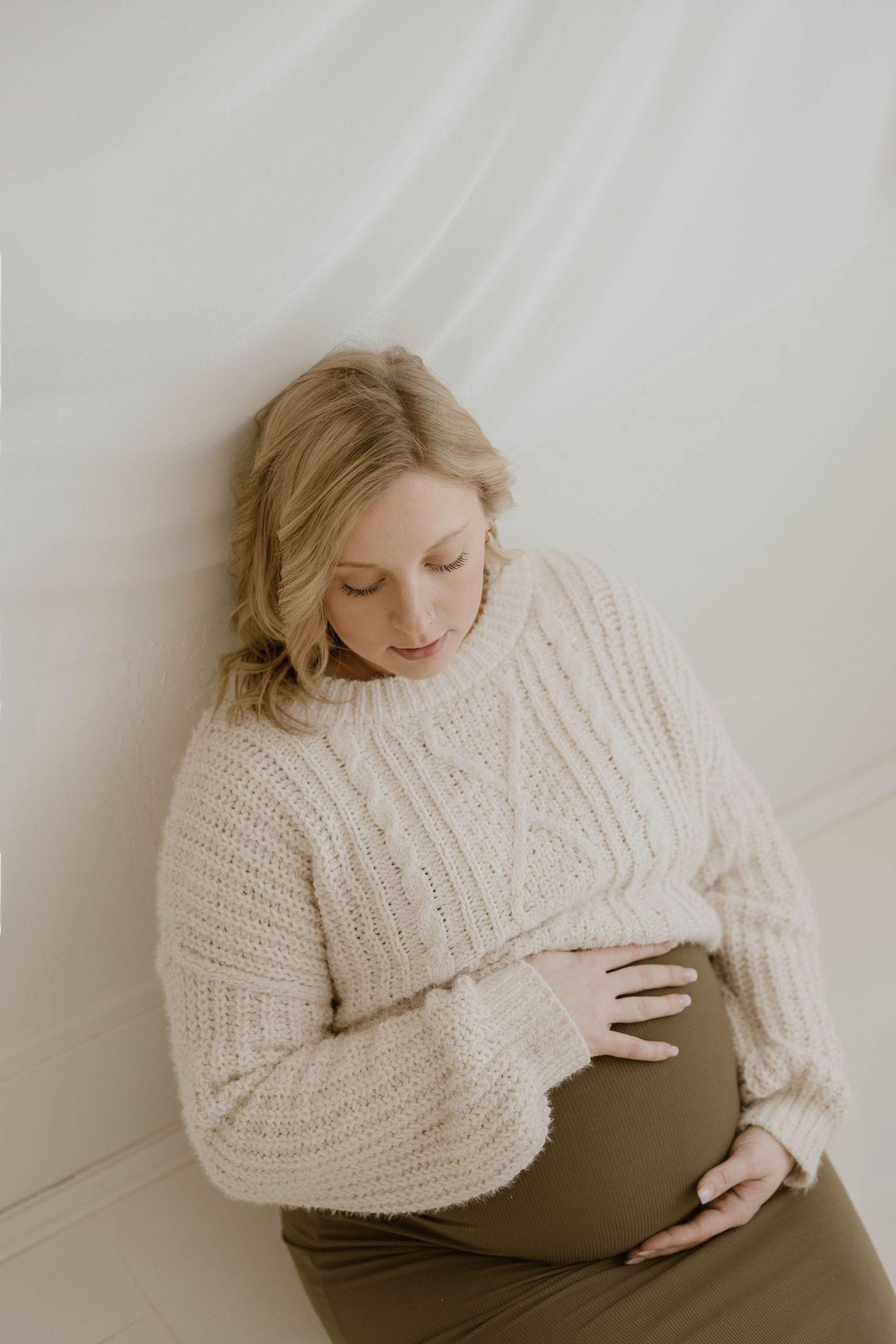 A pregnant woman in a cozy cream knit sweater cradles her belly against a soft white background.