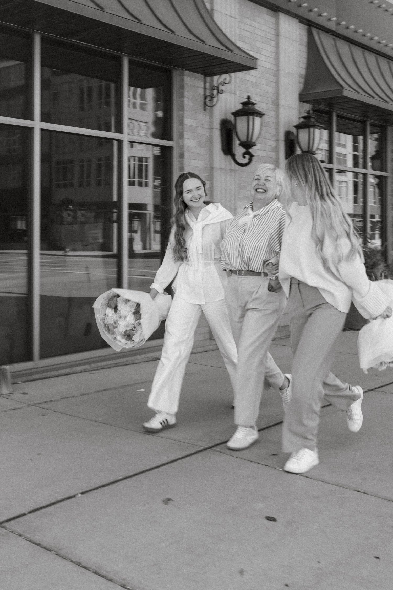 Three women laughing and walking together on a city sidewalk, carrying shopping bags in a black and white photo.