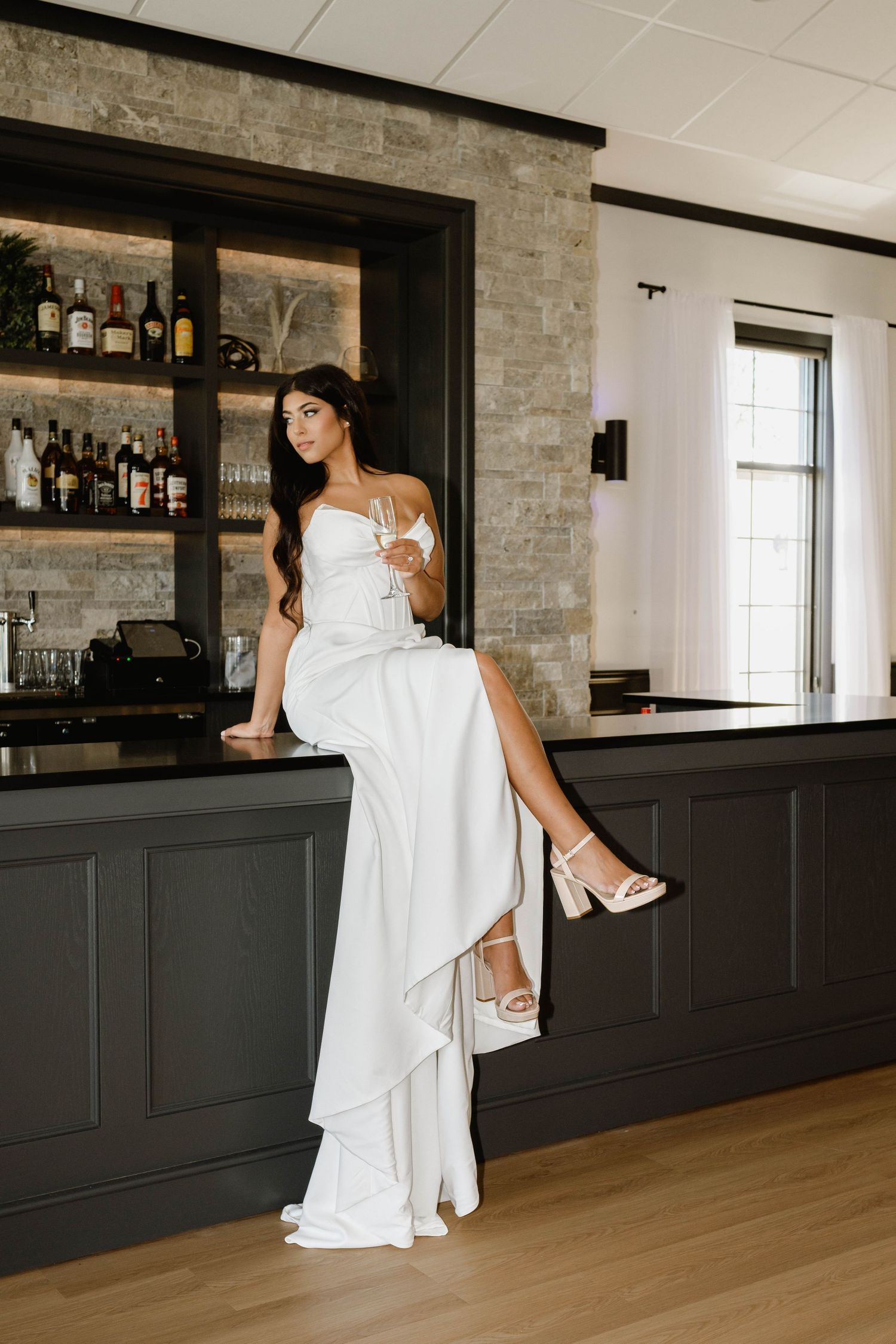 Bride in white gown with slit sits on bar counter holding champagne glass, wearing heels in elegant venue.