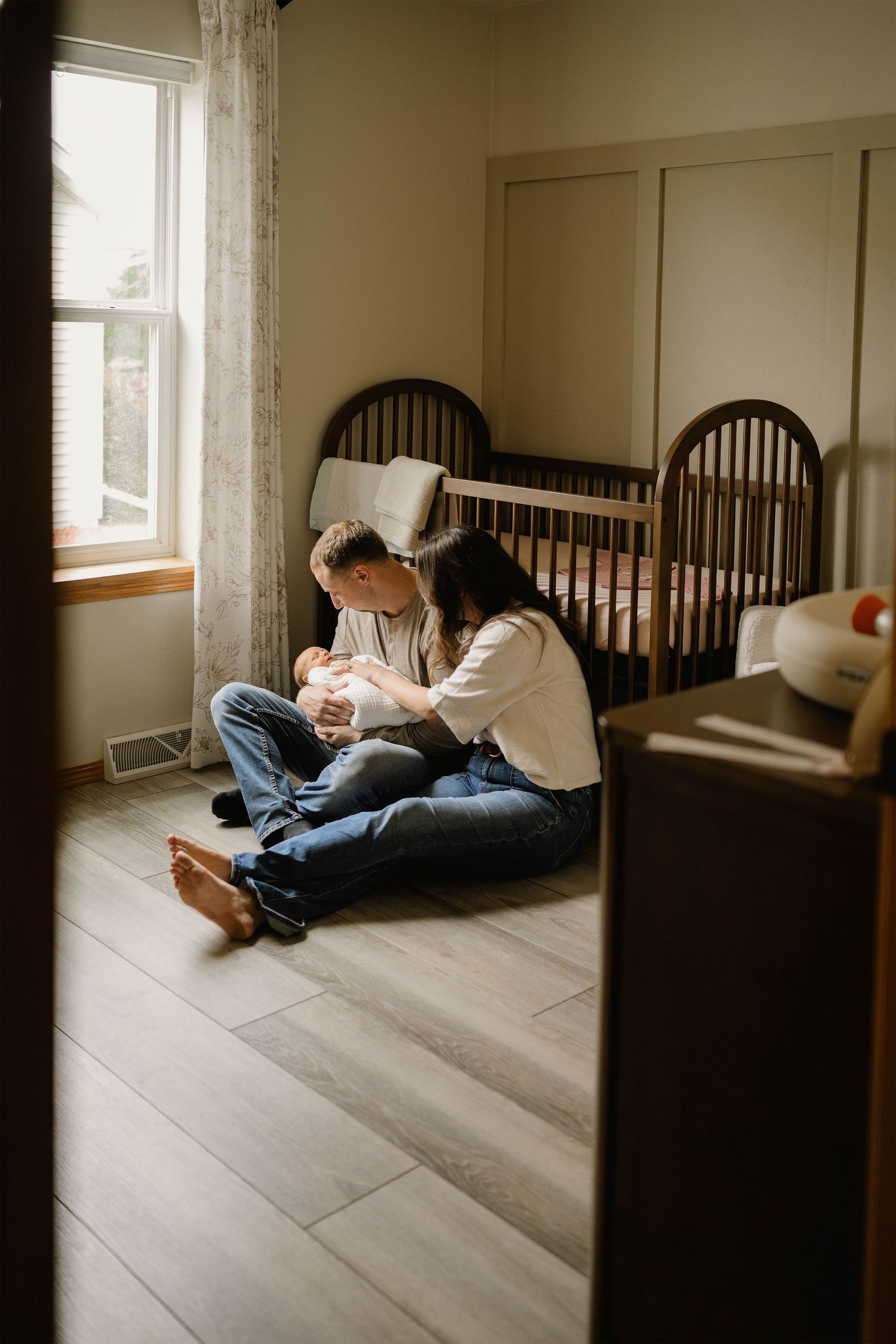 New parents sitting on nursery floor cradling their newborn baby near a crib and window.
