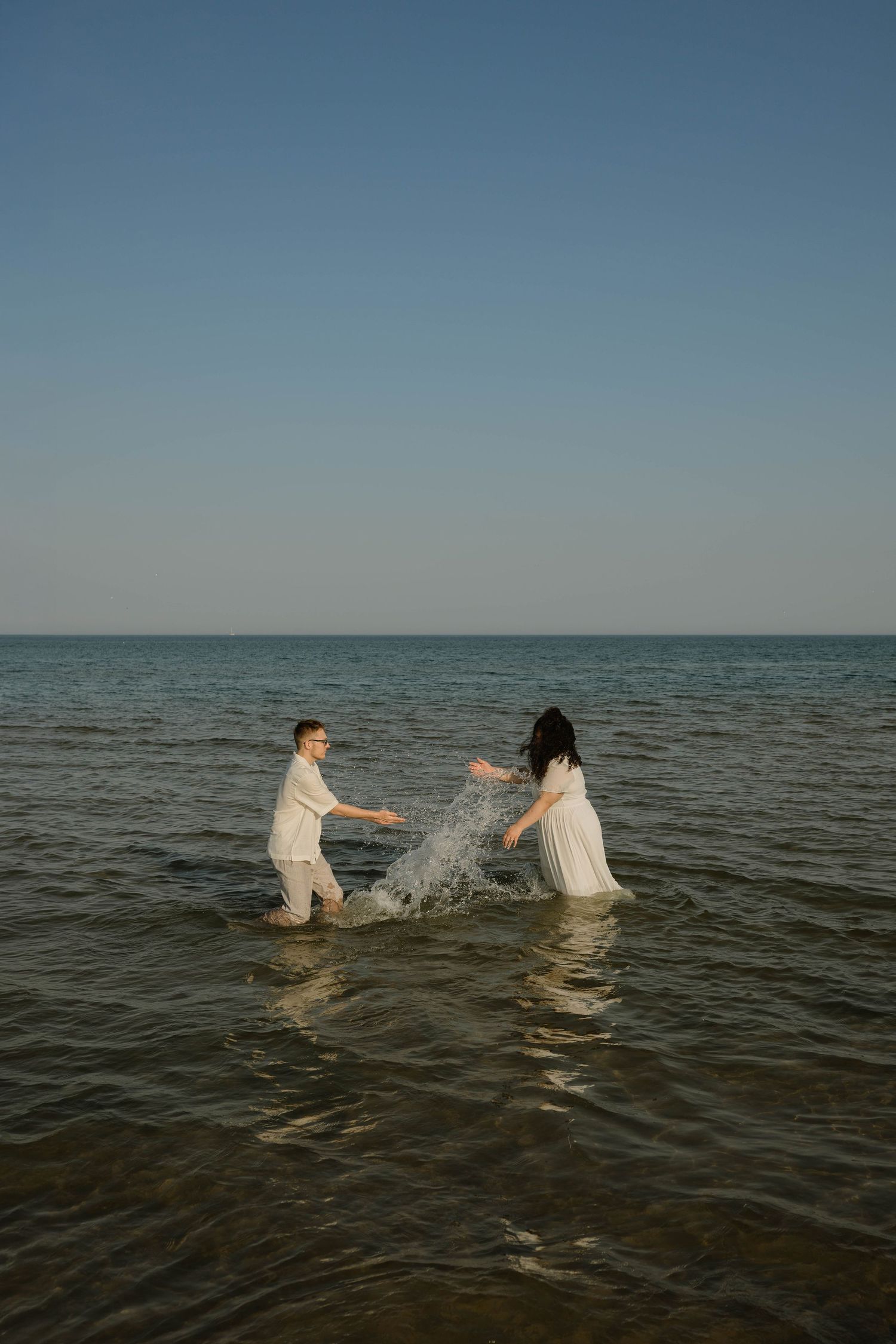 A joyful couple in white outfits splashes in the ocean waves under a clear blue sky.