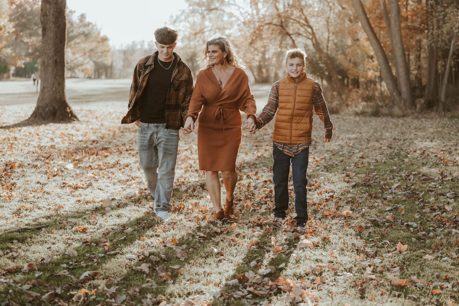 Three people walk together through a fall forest setting wearing coordinating brown and orange toned outfits.
