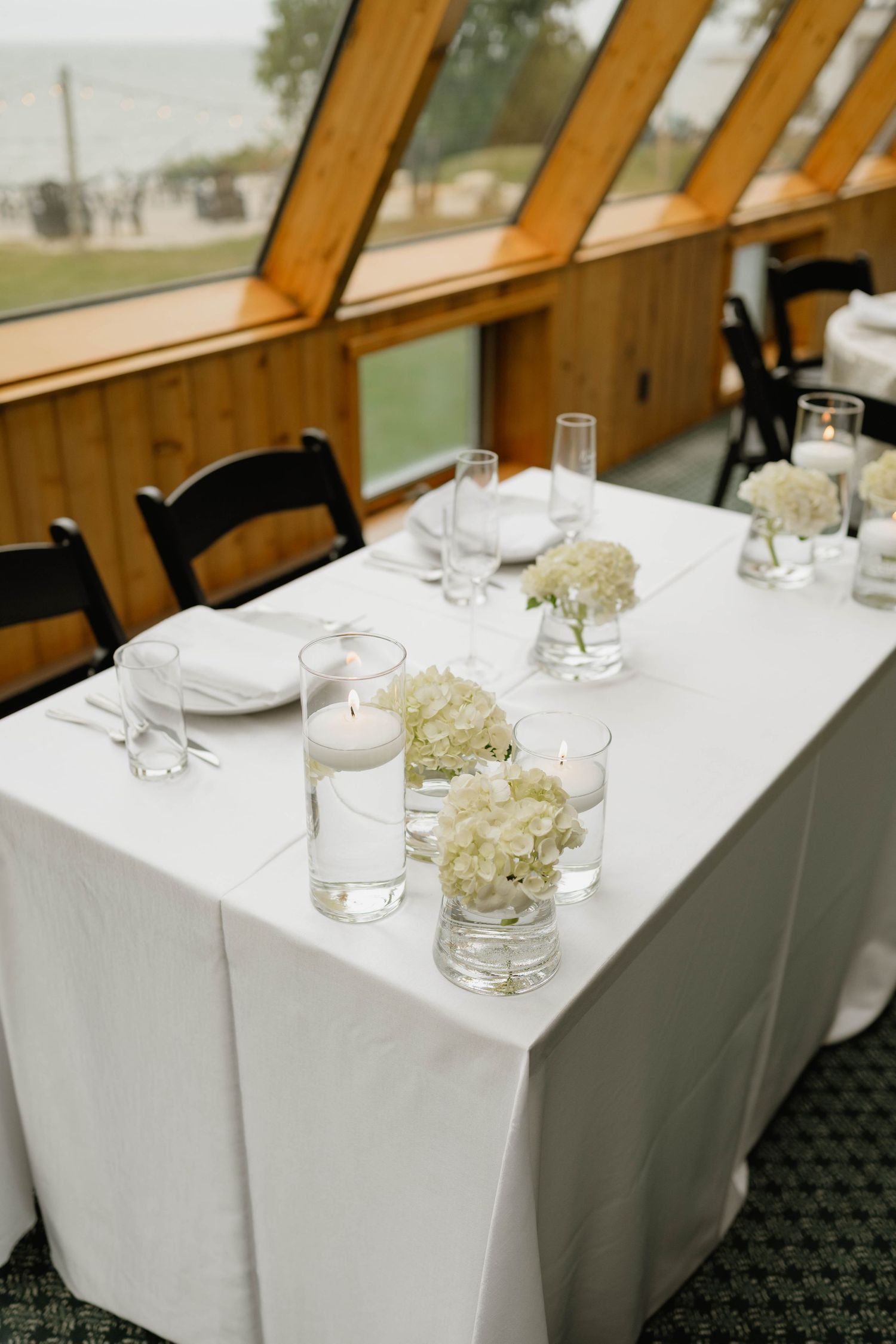 Elegant wedding sweetheart table with white linen, hydrangea centerpieces, floating candles, and black chairs by large windows.