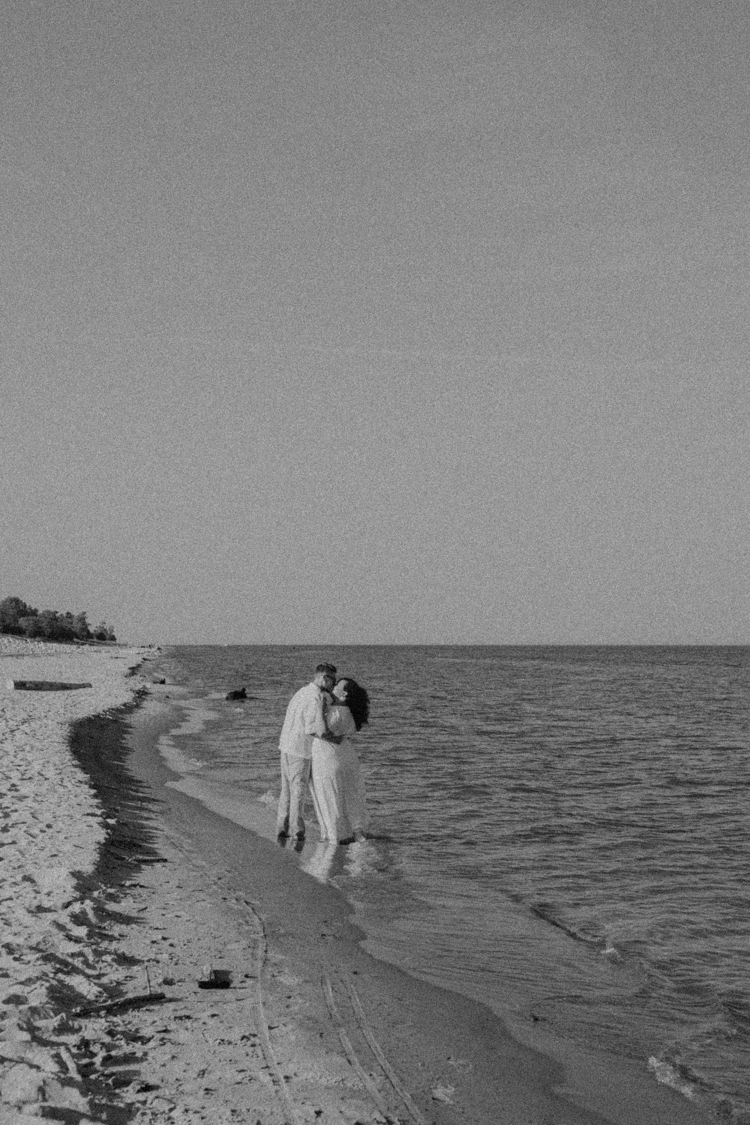 Black and white photo of a couple kissing at the water's edge on a serene beach.