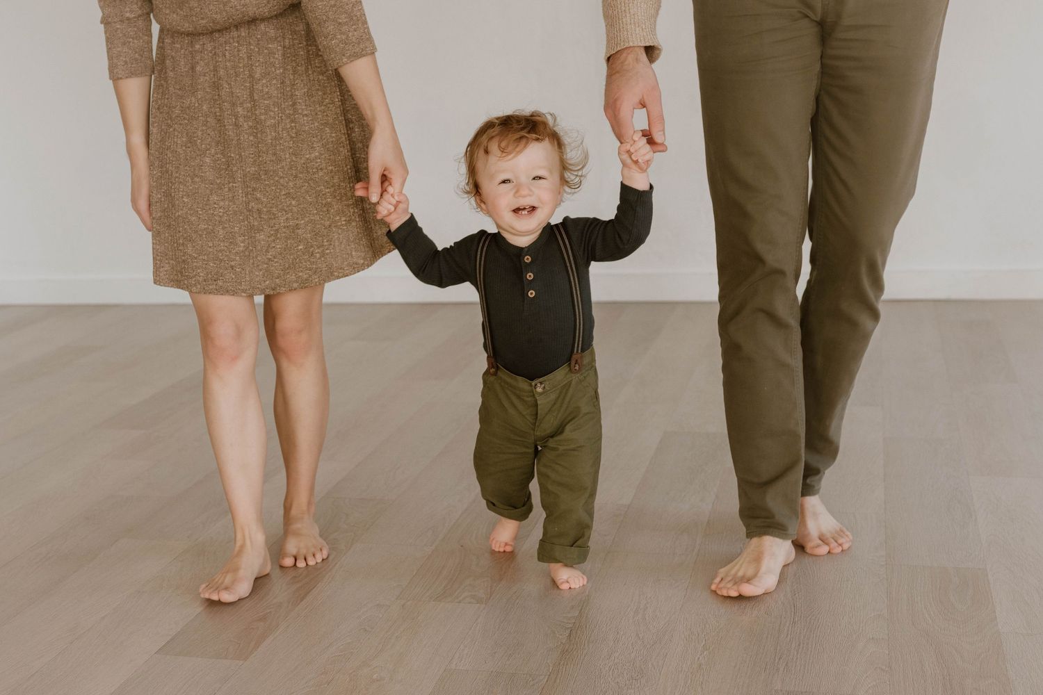 A small child in green overalls holds hands with two adults while standing on hardwood flooring.