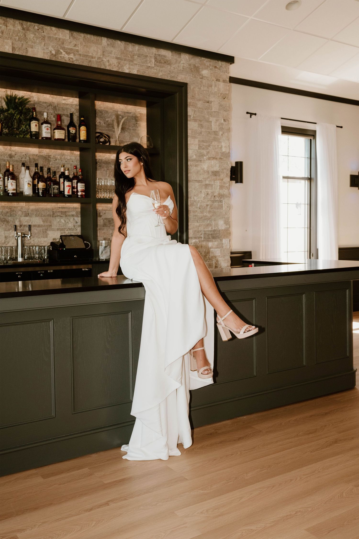 Bride in white gown sitting on bar counter holding champagne glass, elegant wedding venue with dark green cabinetry.