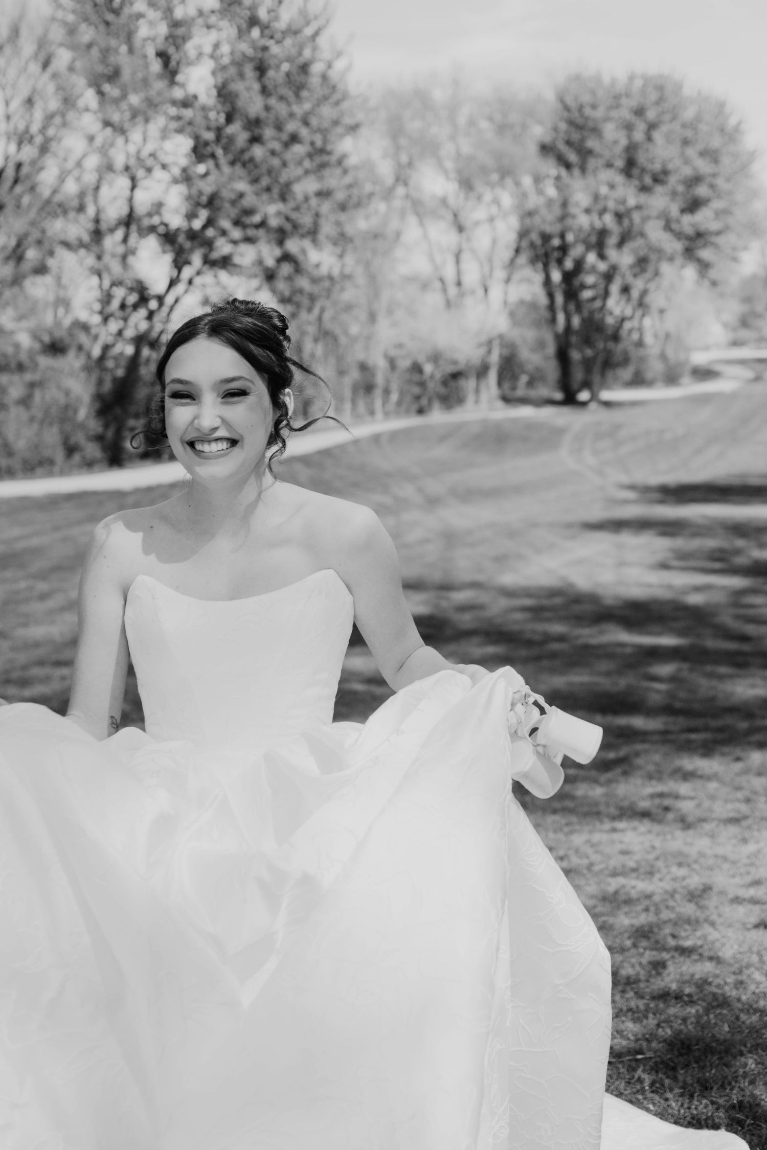Smiling bride in a white ball gown holding her dress while standing outdoors in a black and white photo.