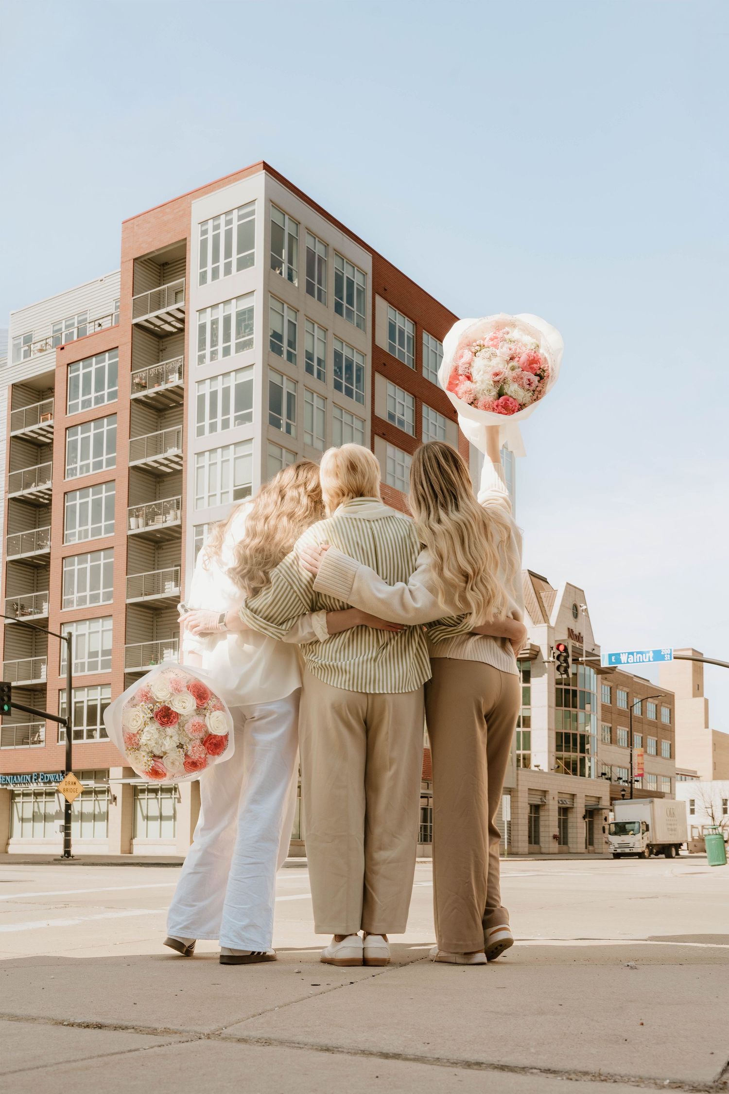 Four women hugging outdoors holding pink floral bouquets with urban buildings in the background.