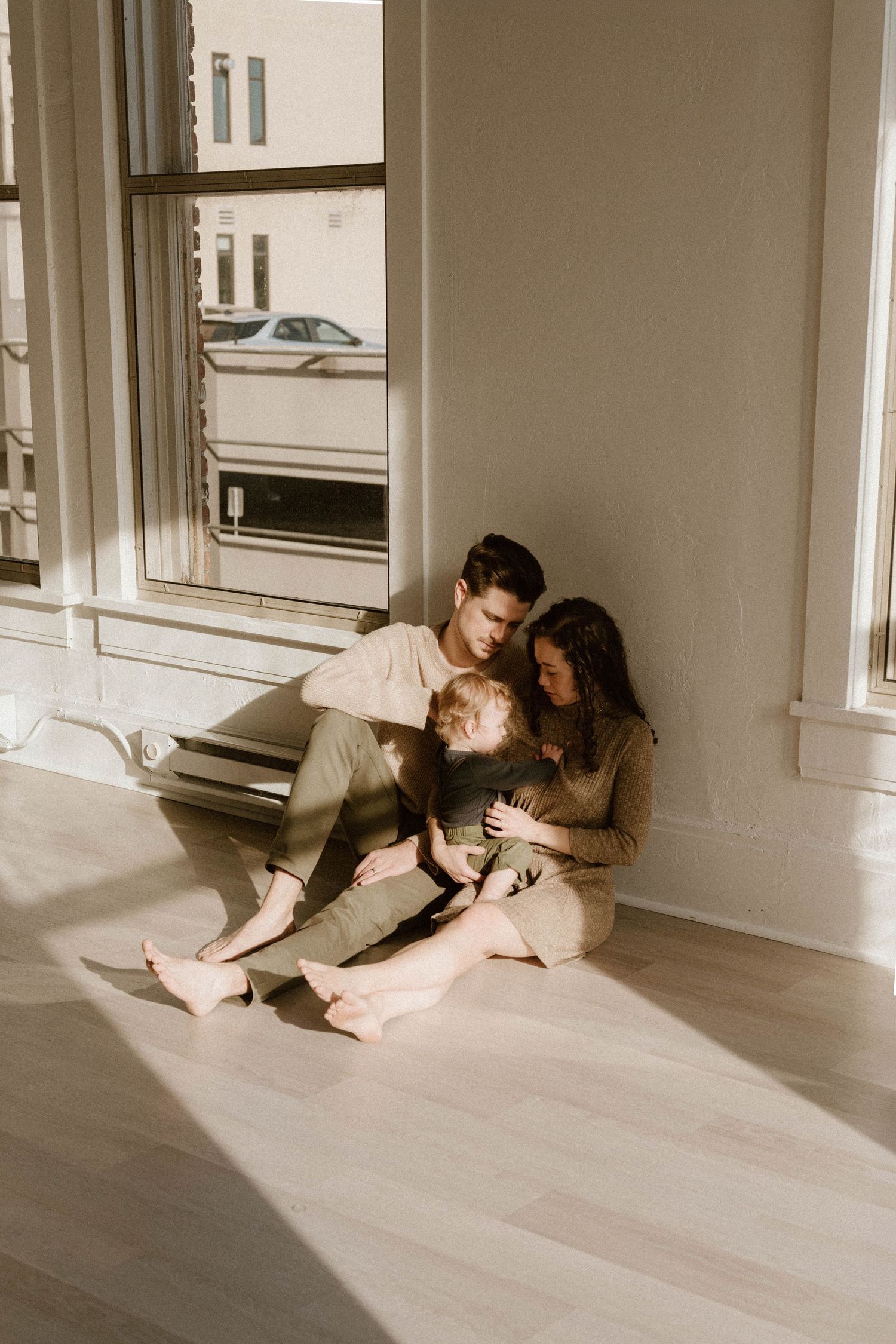 A couple sits together in sunlight on a wooden floor in a minimalist white room, shown in multiple artistic poses.