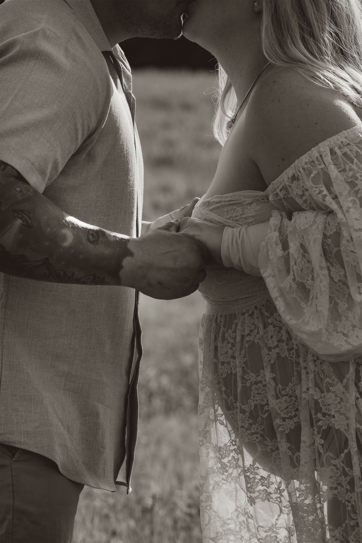 Black and white photo of a tattooed man kissing a pregnant woman in lace dress while holding hands outdoors.