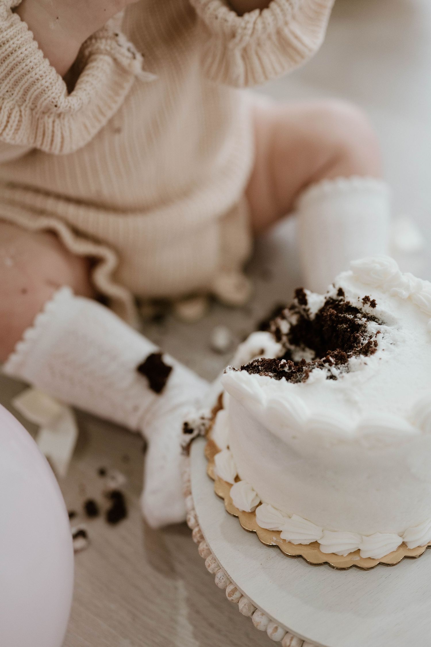 Baby in cream sweater smashing white frosted cake with chocolate crumbles during first birthday cake smash photo session.