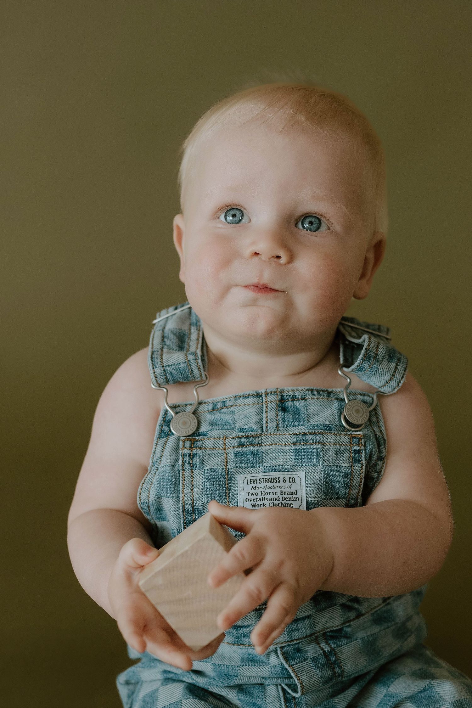Adorable blue-eyed baby in denim overalls holding a wooden block against olive background.