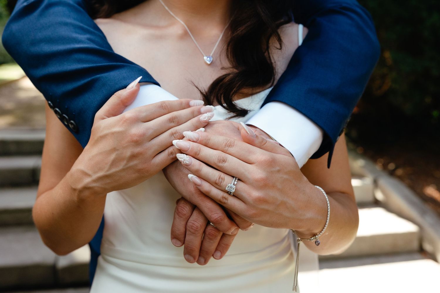 Close-up photo of hands embracing while wearing wedding attire, showing rings and formal sleeves.