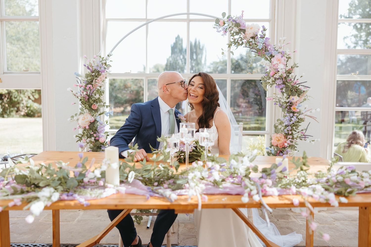 A couple shares a romantic moment at an elegant wedding reception table decorated with pink and purple floral arrangements.