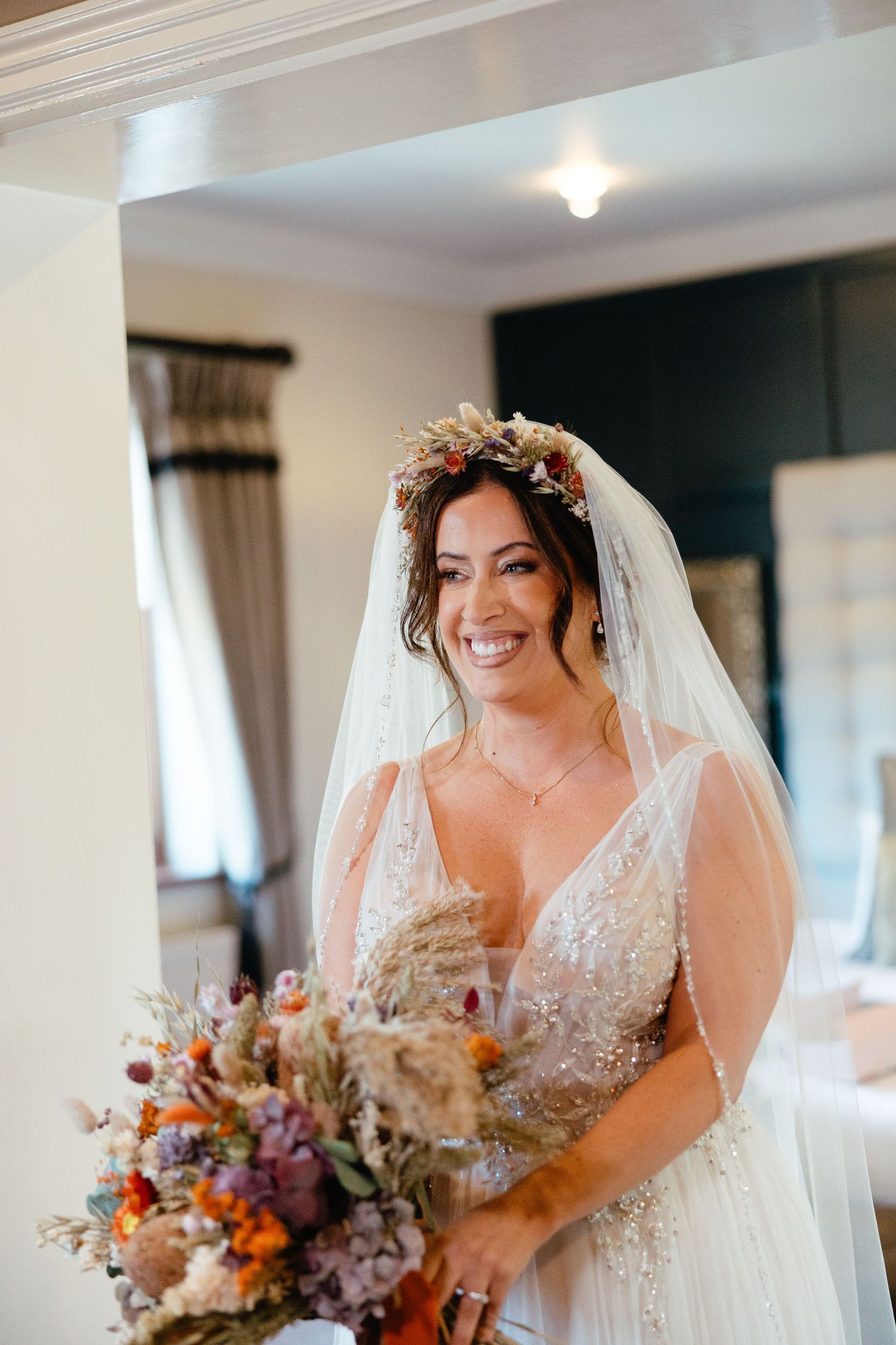A bride in a sparkling white wedding dress holds a vibrant autumn-colored bouquet while smiling indoors.