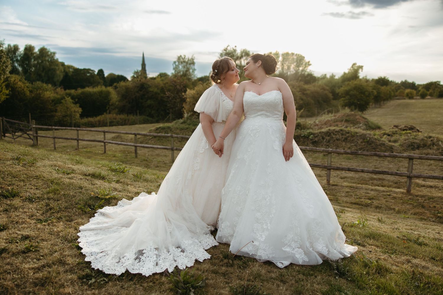 Two white wedding gowns flowing in the wind against a scenic rural landscape at sunset.