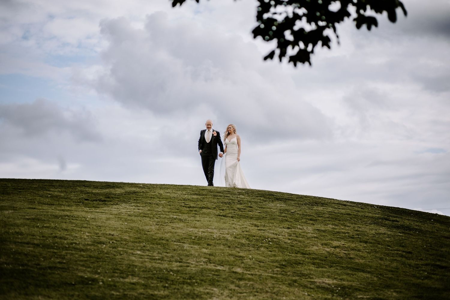 Silhouettes of a couple walk along a grassy hilltop against a dramatic cloudy sky during wedding photos.