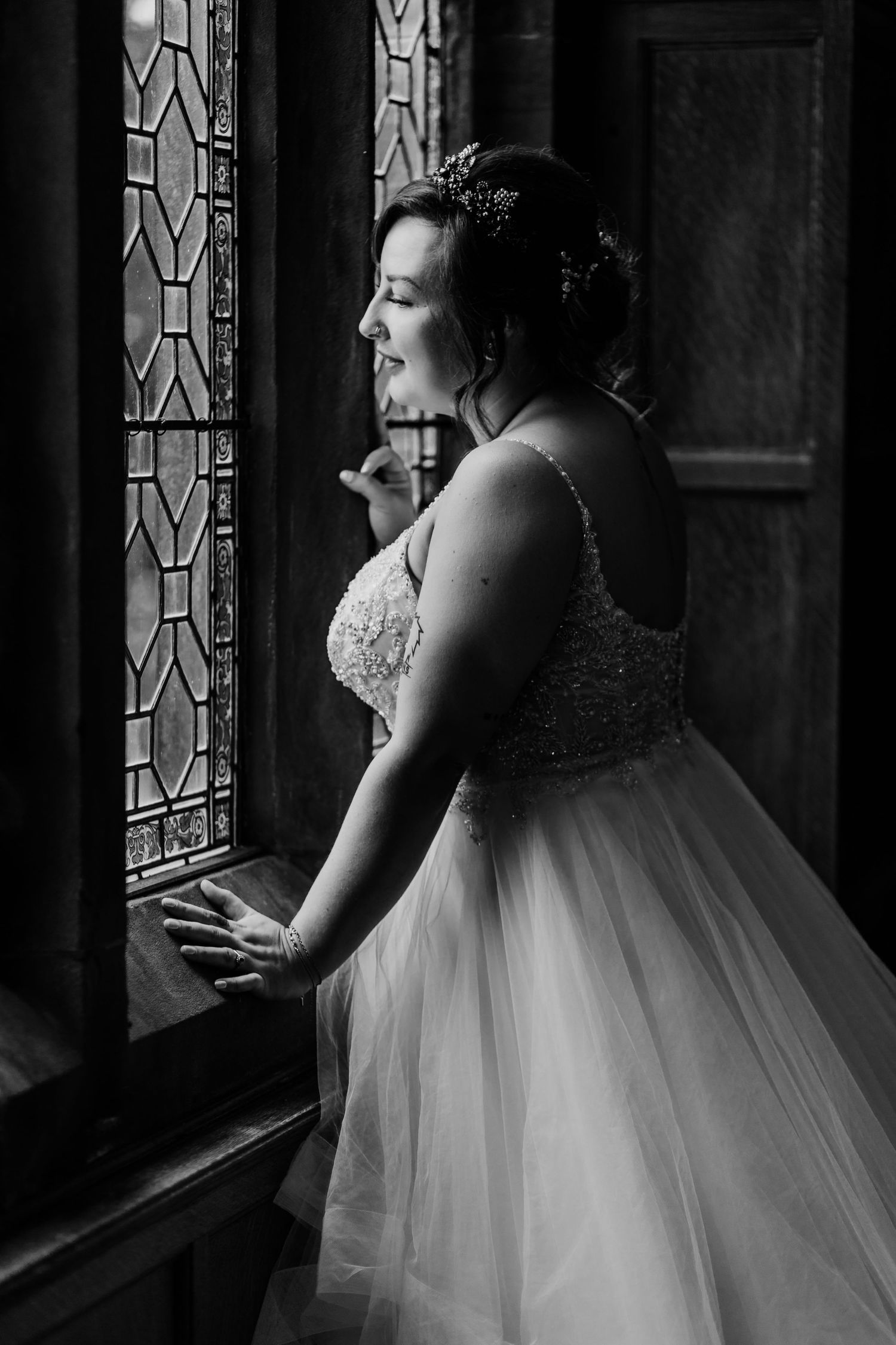 Black and white portrait of a bride in a full tulle gown gazing through an ornate window in dramatic lighting.